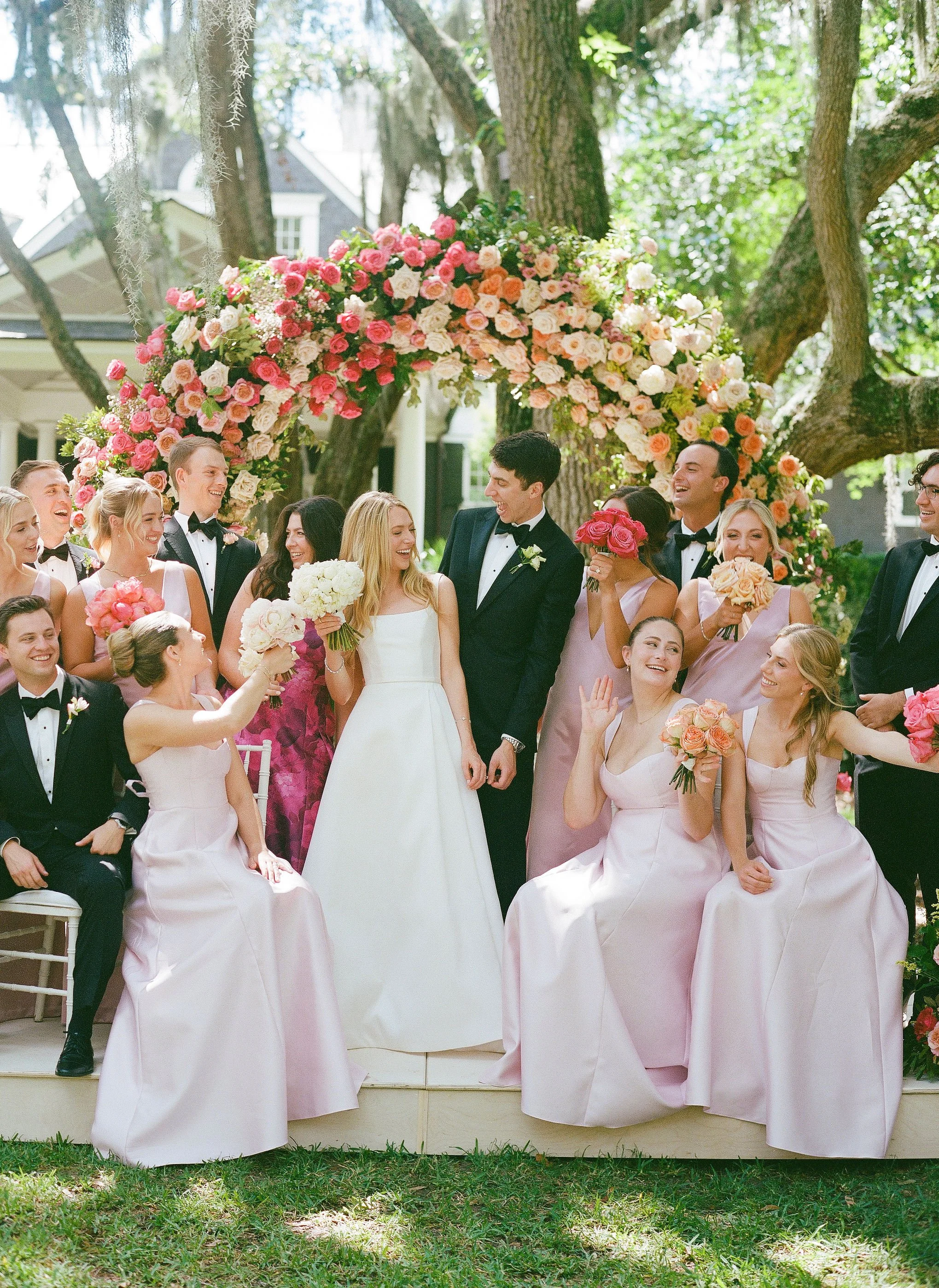 Bride, groom, bridesmaids, and groomsmen gathered beneath a colorful floral arch at an outdoor Charleston wedding.