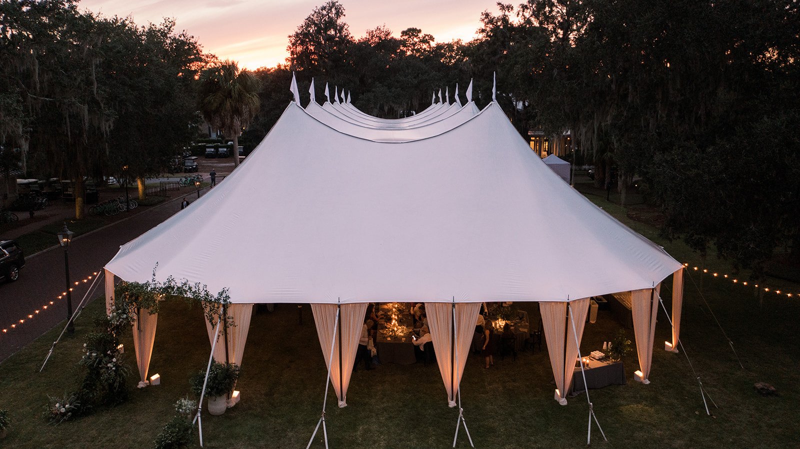 Aerial view of a large white sailcloth wedding tent illuminated at dusk beneath surrounding trees.