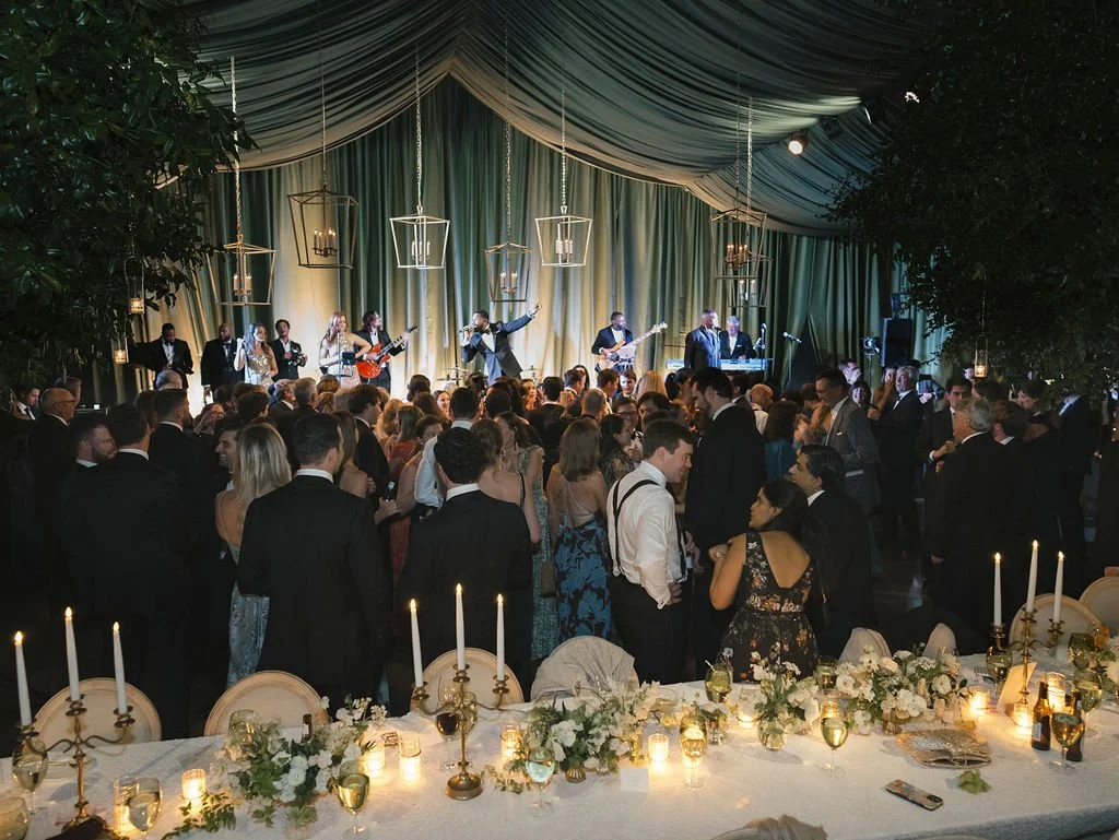 Wedding guests dancing to a live band inside a green-draped tent with candlelit tables nearby.