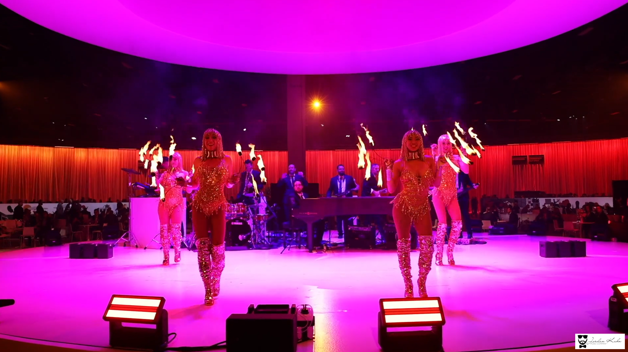 Fire performers in gold costumes perform in front of a live wedding band during an energetic reception illuminated with pink and red lighting.