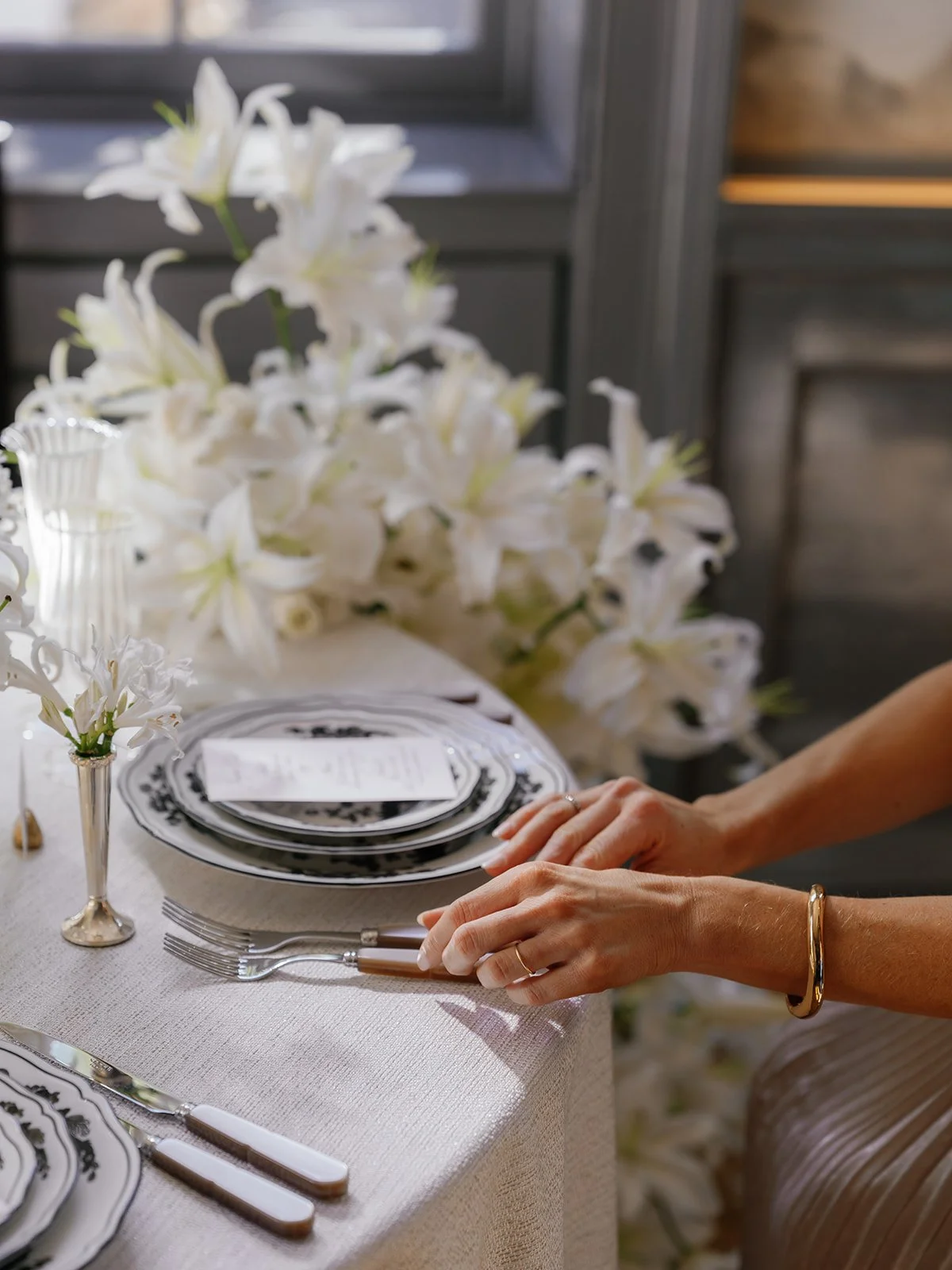 Close-up of hands adjusting flatware beside patterned china and white lily arrangements on a softly textured ivory tablecloth.
