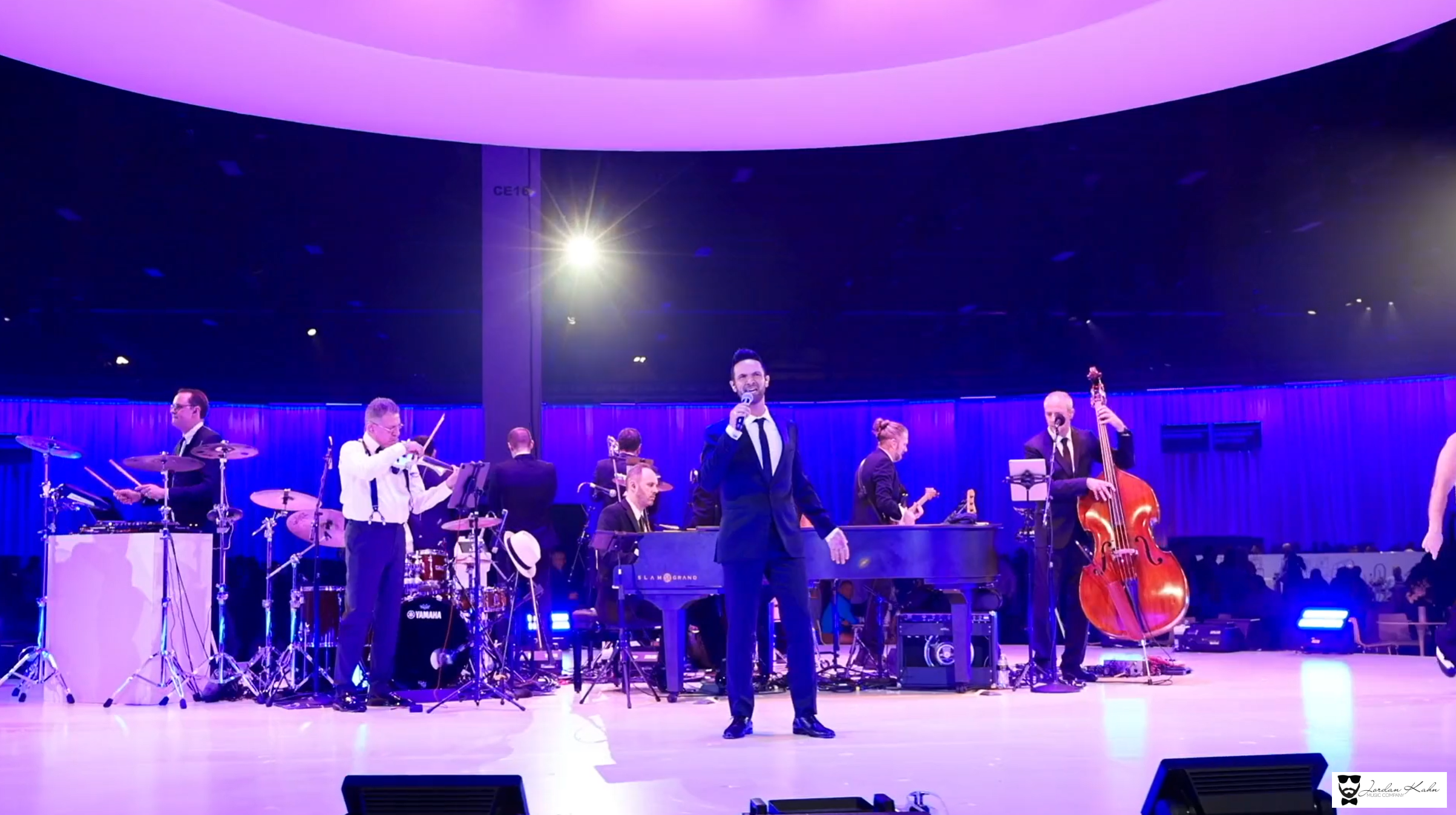 Jordan Kahn Music Company performs live at a wedding reception with a vocalist, violinist, drummer, pianist, guitarist, and upright bassist under blue and purple lighting.