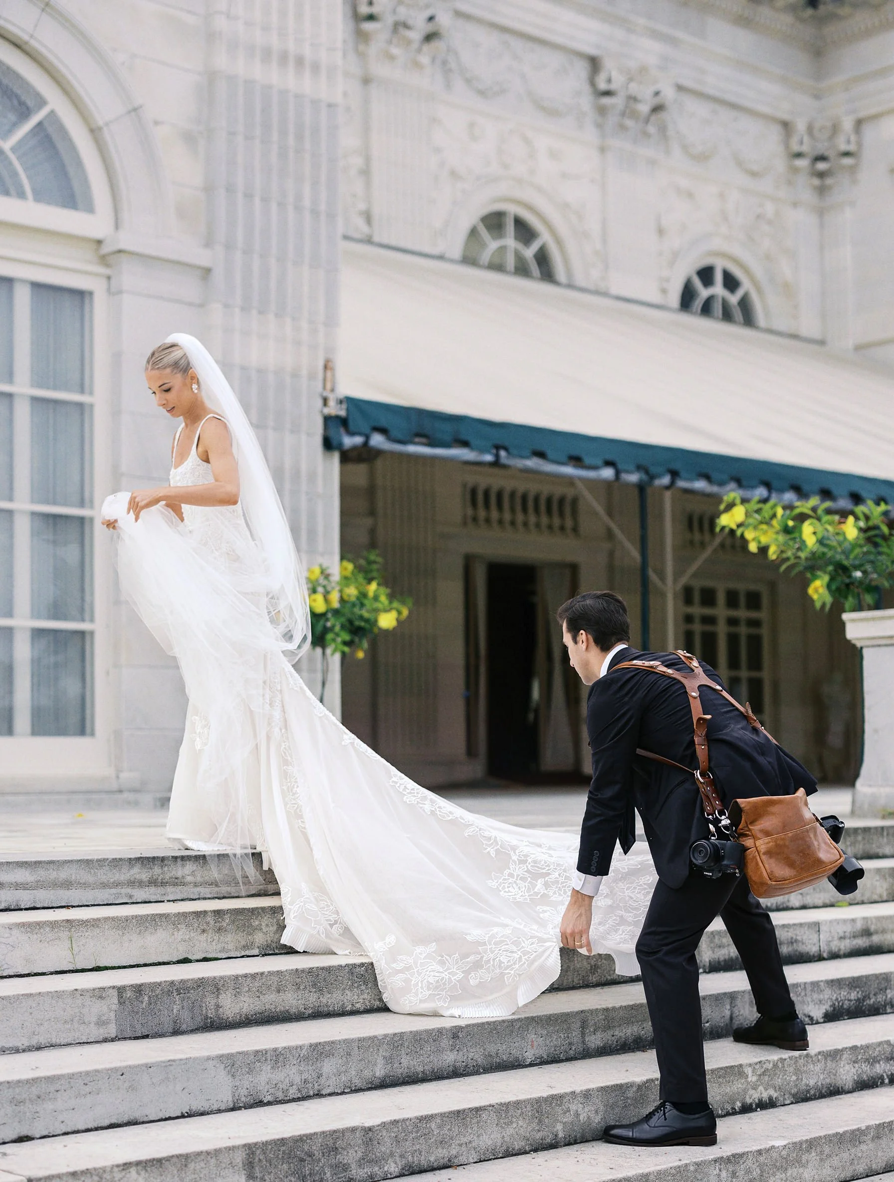 Bride in a lace gown and long veil steps up stone stairs outside a grand building while a photographer in a suit adjusts the train.