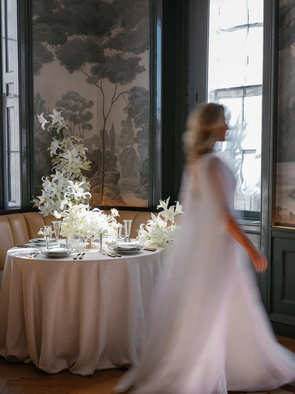 A bride moves past a linen-draped reception table with white lilies, crystal glassware, and layered place settings.