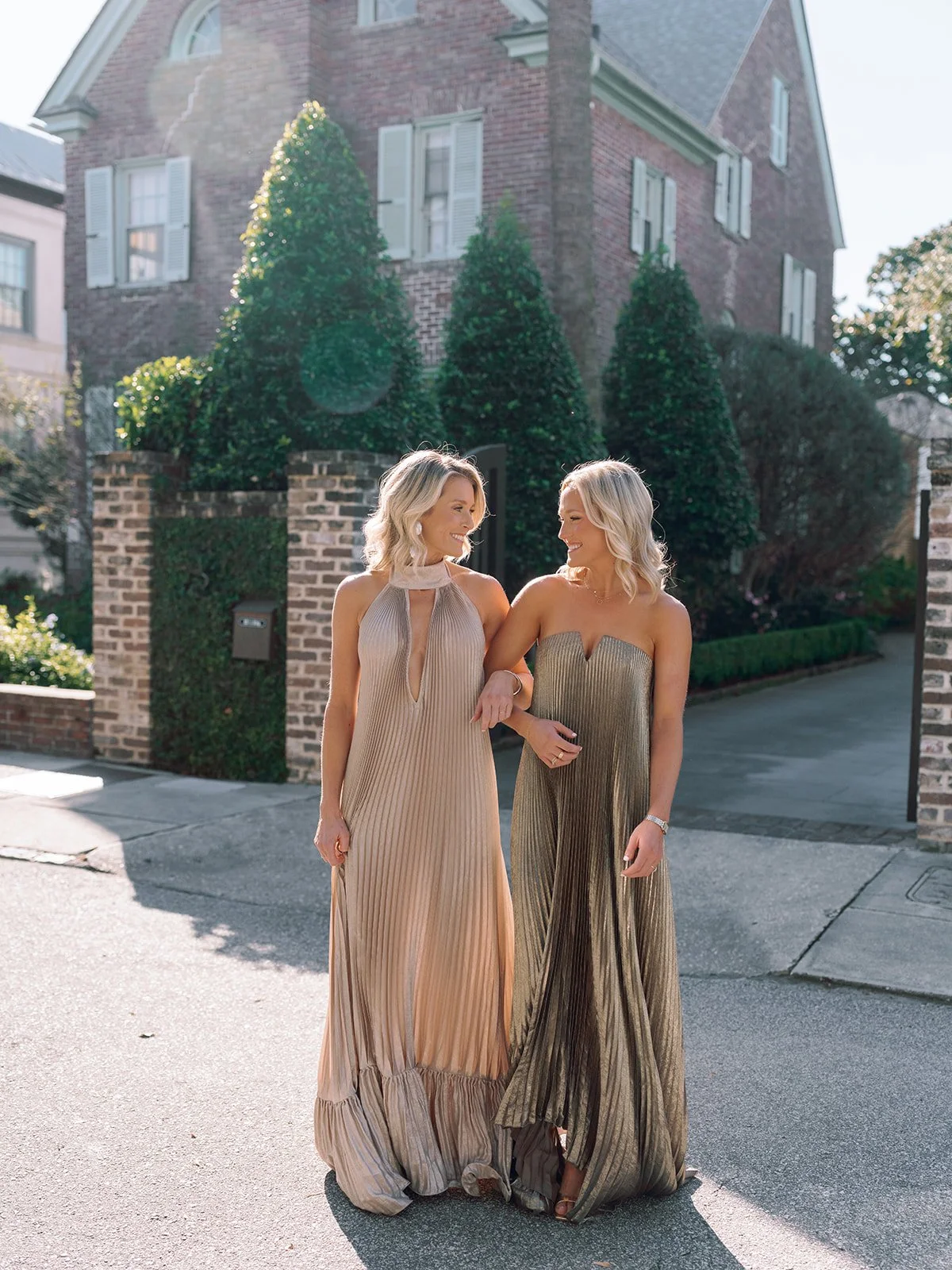 Two women in metallic gowns walk arm-in-arm on a sunny Charleston street in front of a historic brick home and manicured greenery.