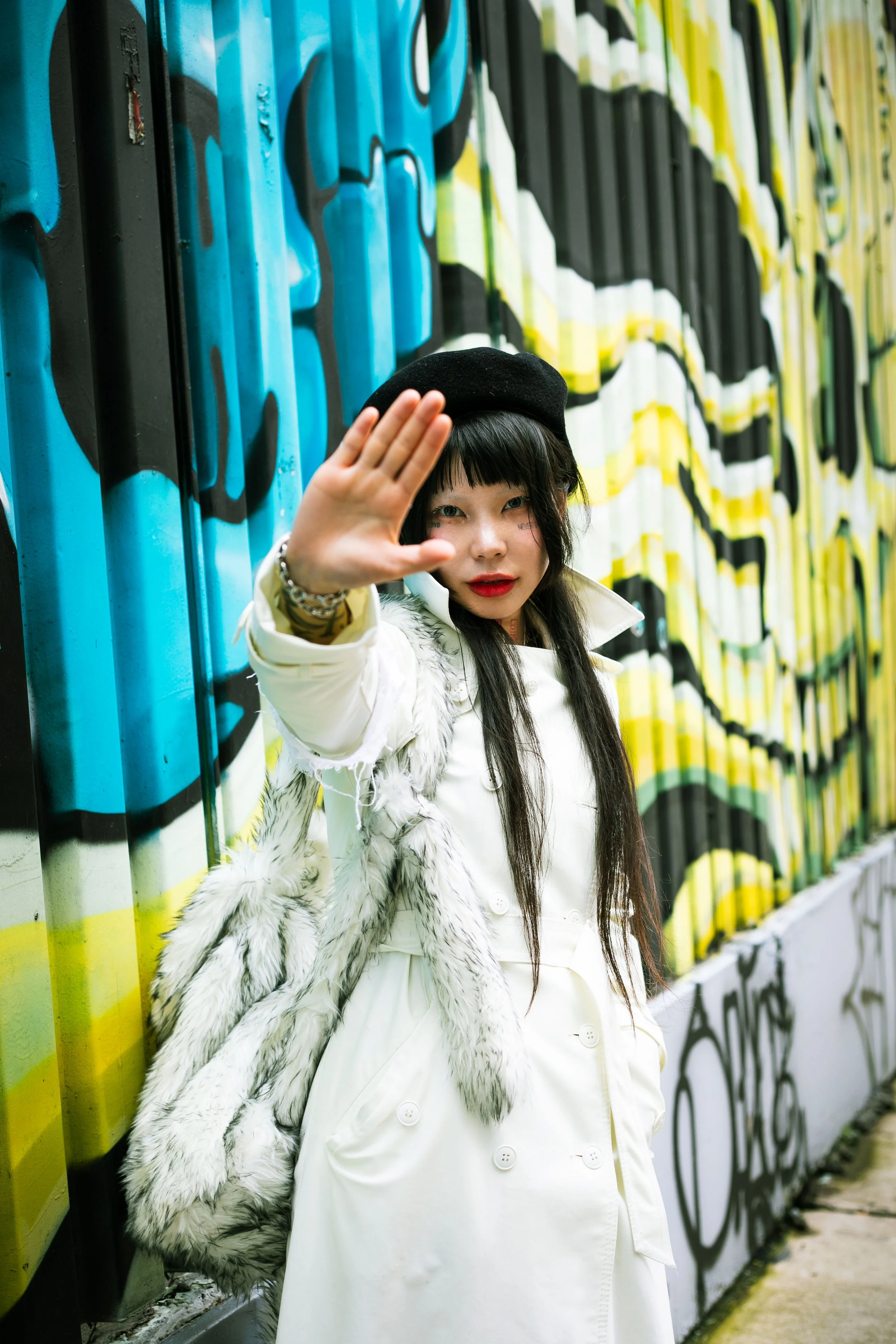 Korean woman with long straight black hair wearing a black beret and white trench coat. She stands in front of an outdoor wall painted blue, yellow, and red, reaching her hand out towards the camera.