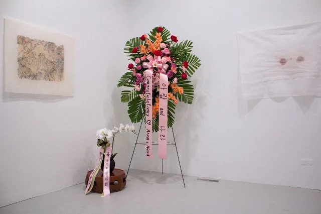 Corner of a white room with a congratulatory garland at the center with yellow, red, and pink flowers and ribbons that read "Aram and Nara." Two white and ivory fabric paintings hang on the left and right walls.