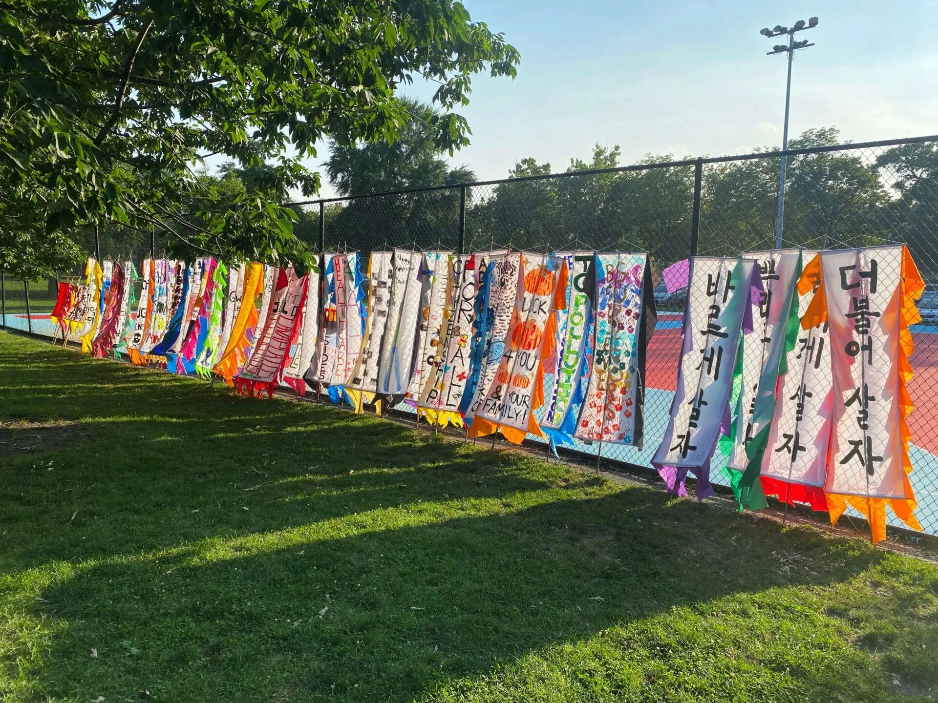 Around twenty vertically long fabric banners hang on a wired fence. They shed long shadows on the green pasture in front of them, and a part of a tree partially obscures the end of the row of banners.