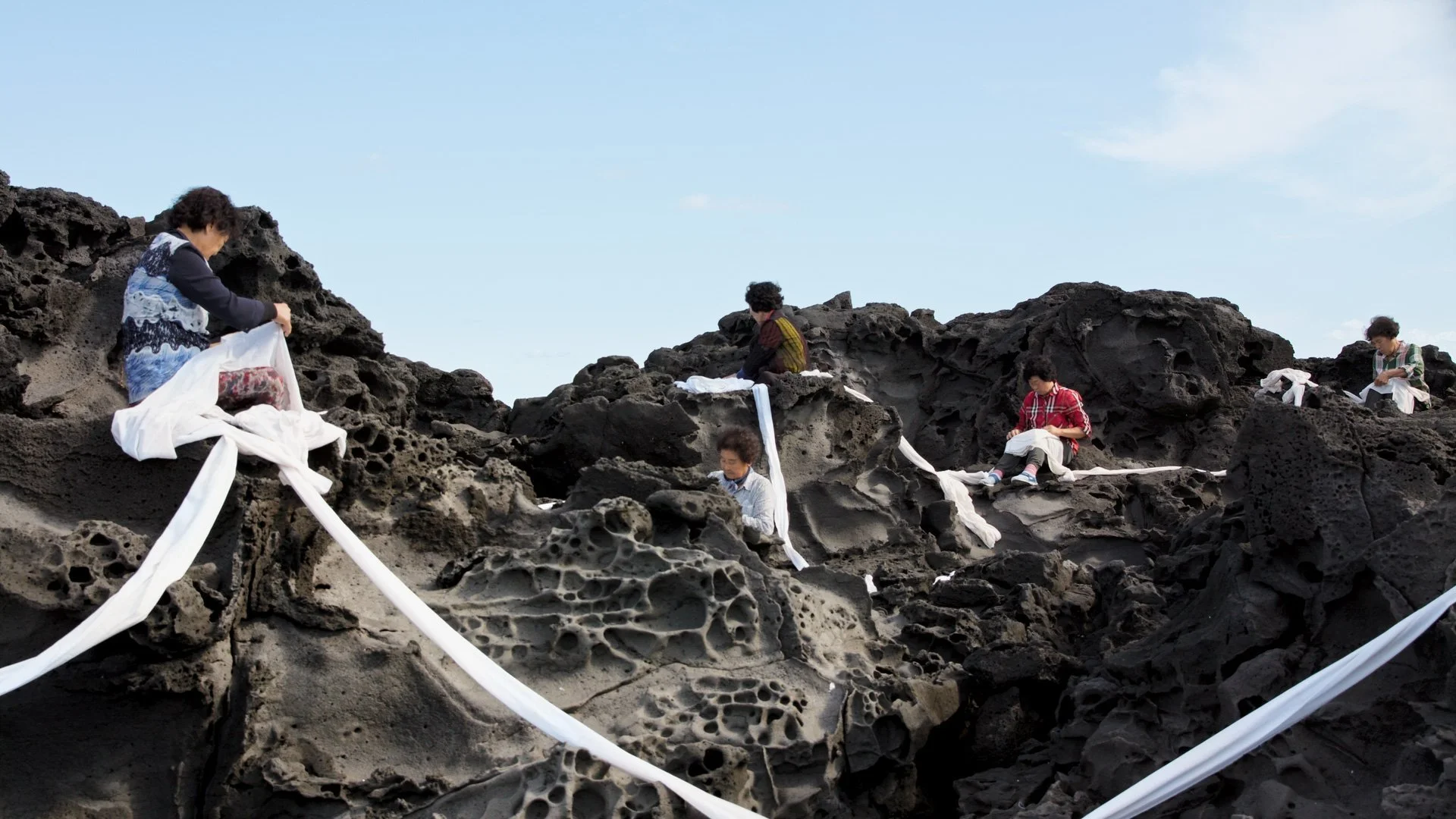 Still of a Jain Jin Kaisen's video work, of four elderly women sitting on a shore of black and grey, porous rocks. They sit at different spots of the shore, holding a long white strand of cloth that continues between them.
