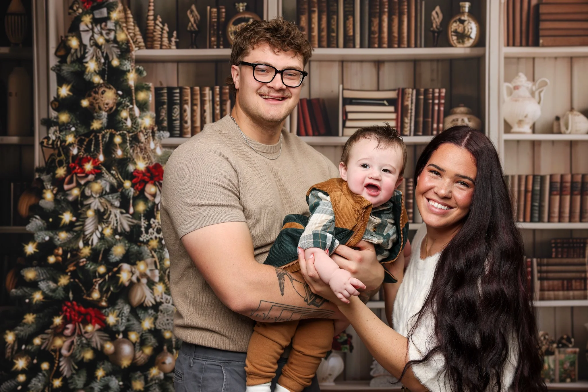 Professional Christmas studio family portrait featuring a smiling mother, father, and toddler boy posed in front of a decorated Christmas tree by J. Warrington Visual Creations in Findlay, Ohio.