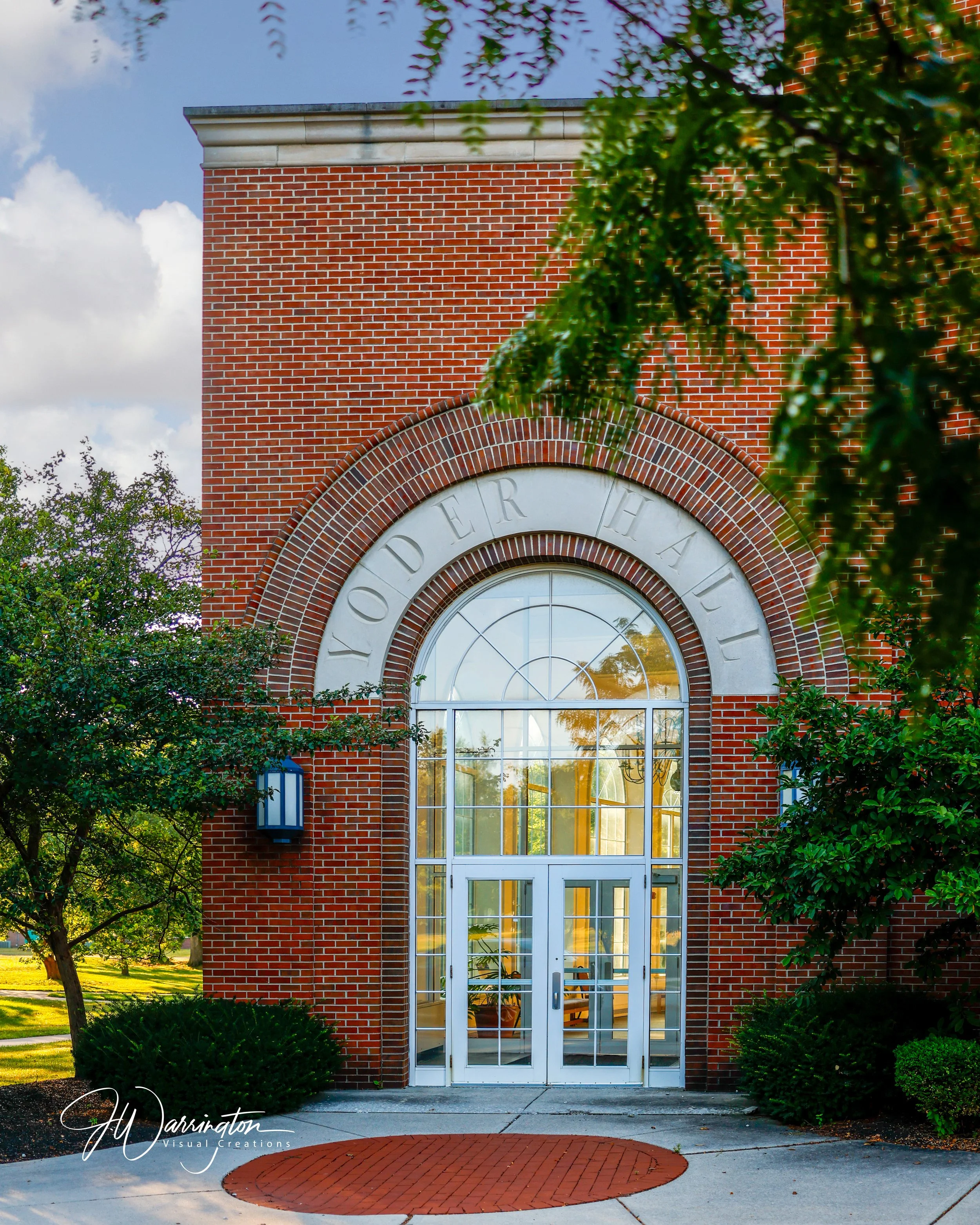 A brick building with a large arched window, partially obscured by trees and shrubs, under a partly cloudy sky at Bluffton University.
