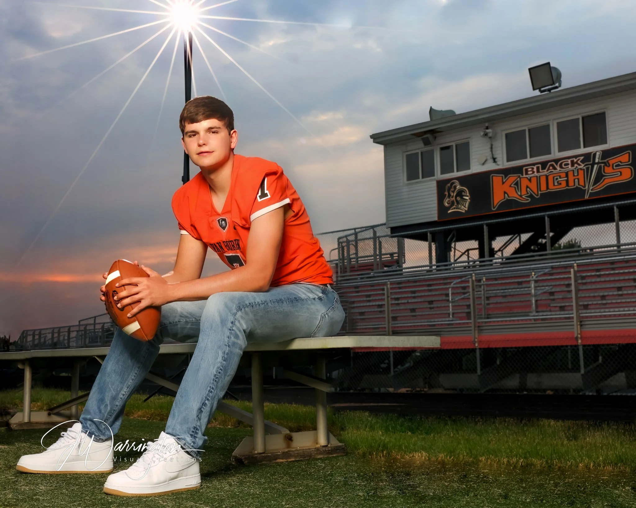 A young man in an orange football jersey and jeans sits on a bench holding a football. The background shows a stadium with a sign that reads "Black Knights" and bleachers, under a cloudy sky by northwest ohio photographer