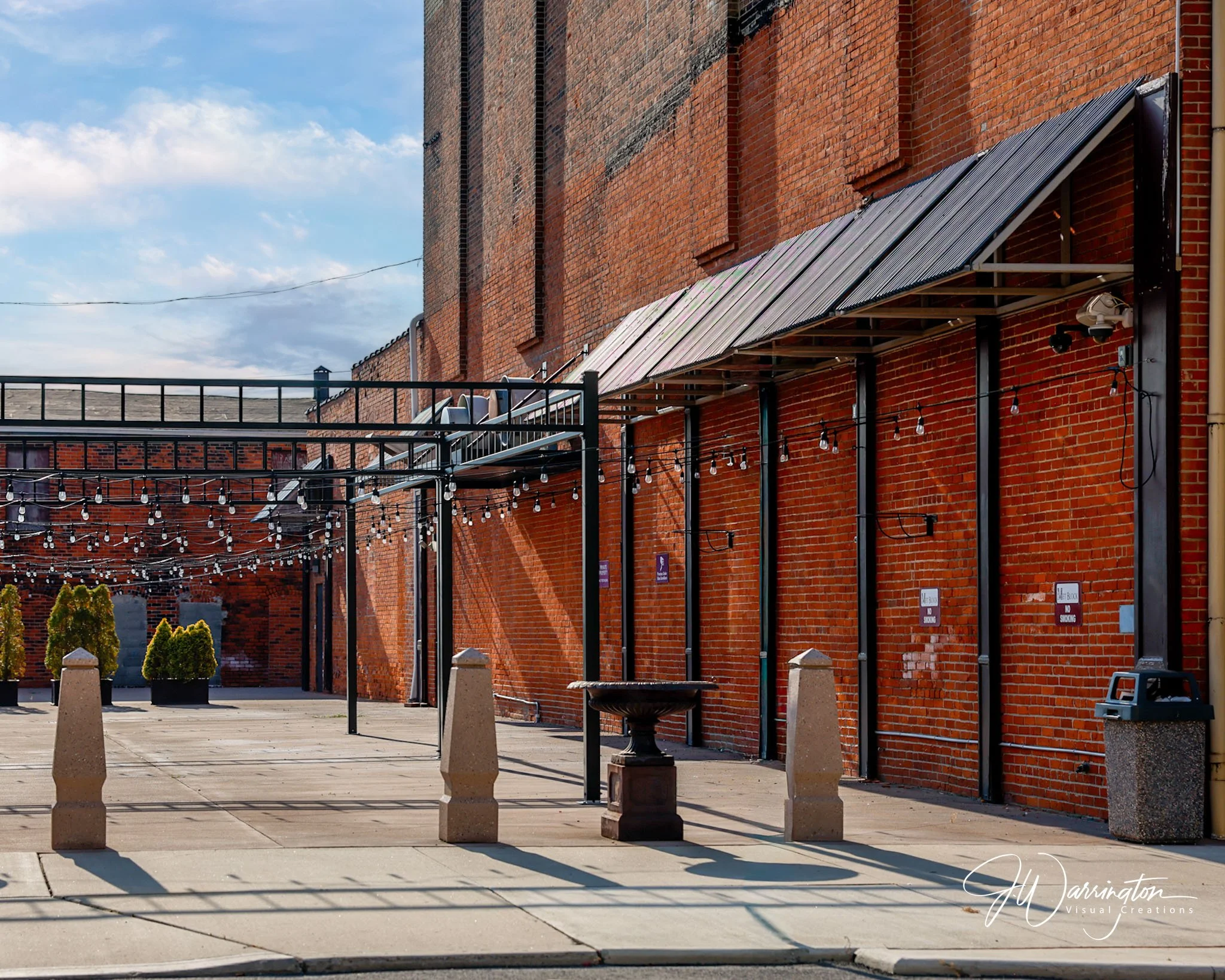 Red brick building in Downtown Lima with a metal awning above outdoor seating and string lights hanging nearby. Several small bushes are in front of the building, with concrete bollards separating the sidewalk from the seating area.