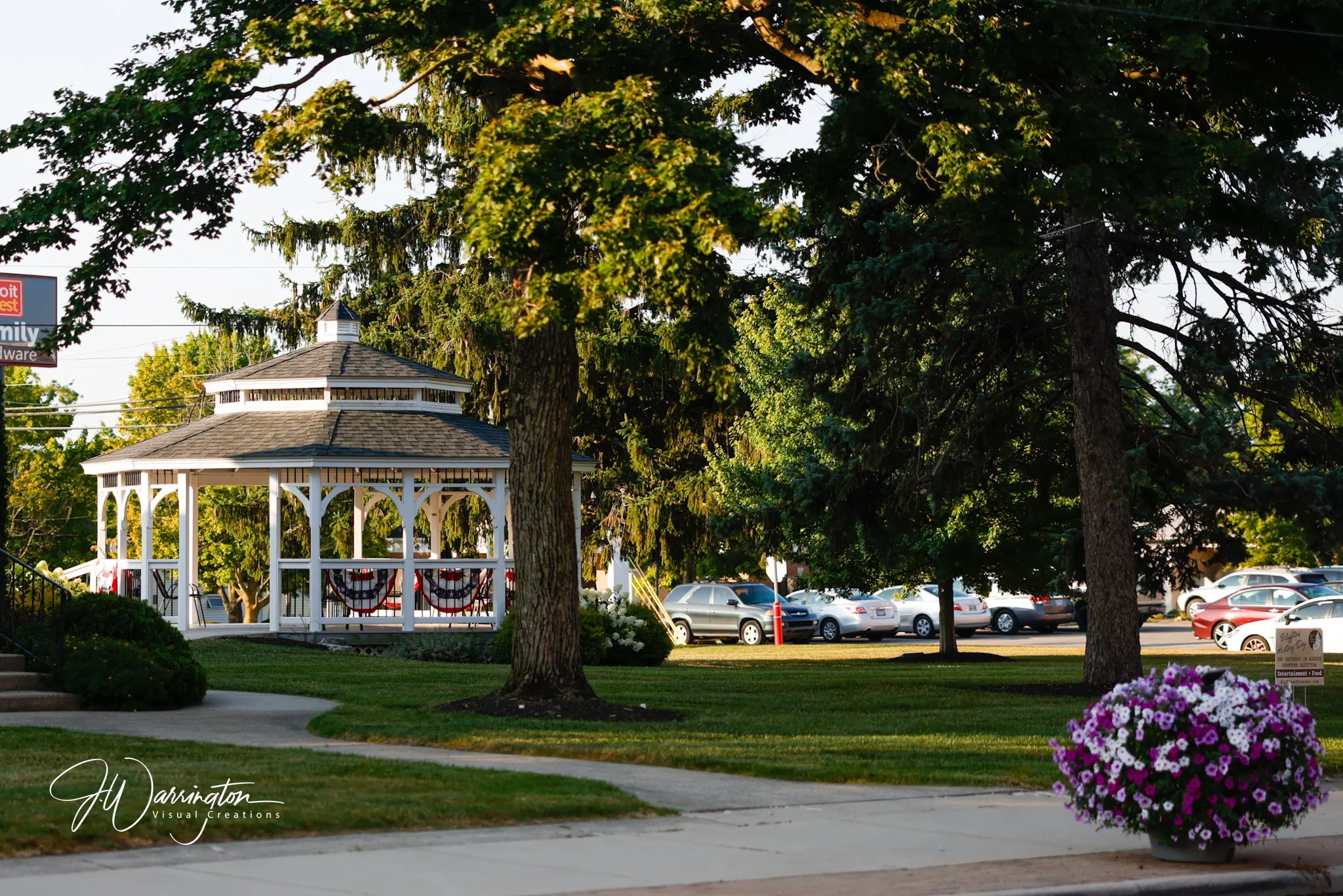 A gazebo in Downtown Bluffton, decorated in with patriotic bunting in a park with trees, parked cars, and purple flowers nearby.