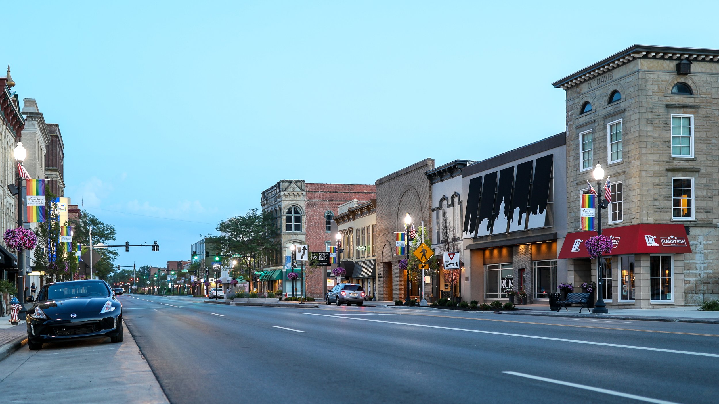 A downtown street scene during early evening, featuring historic buildings, cars parked along the street, and American flags and rainbow flags hanging from lamp posts, with flowering baskets decorating the area.