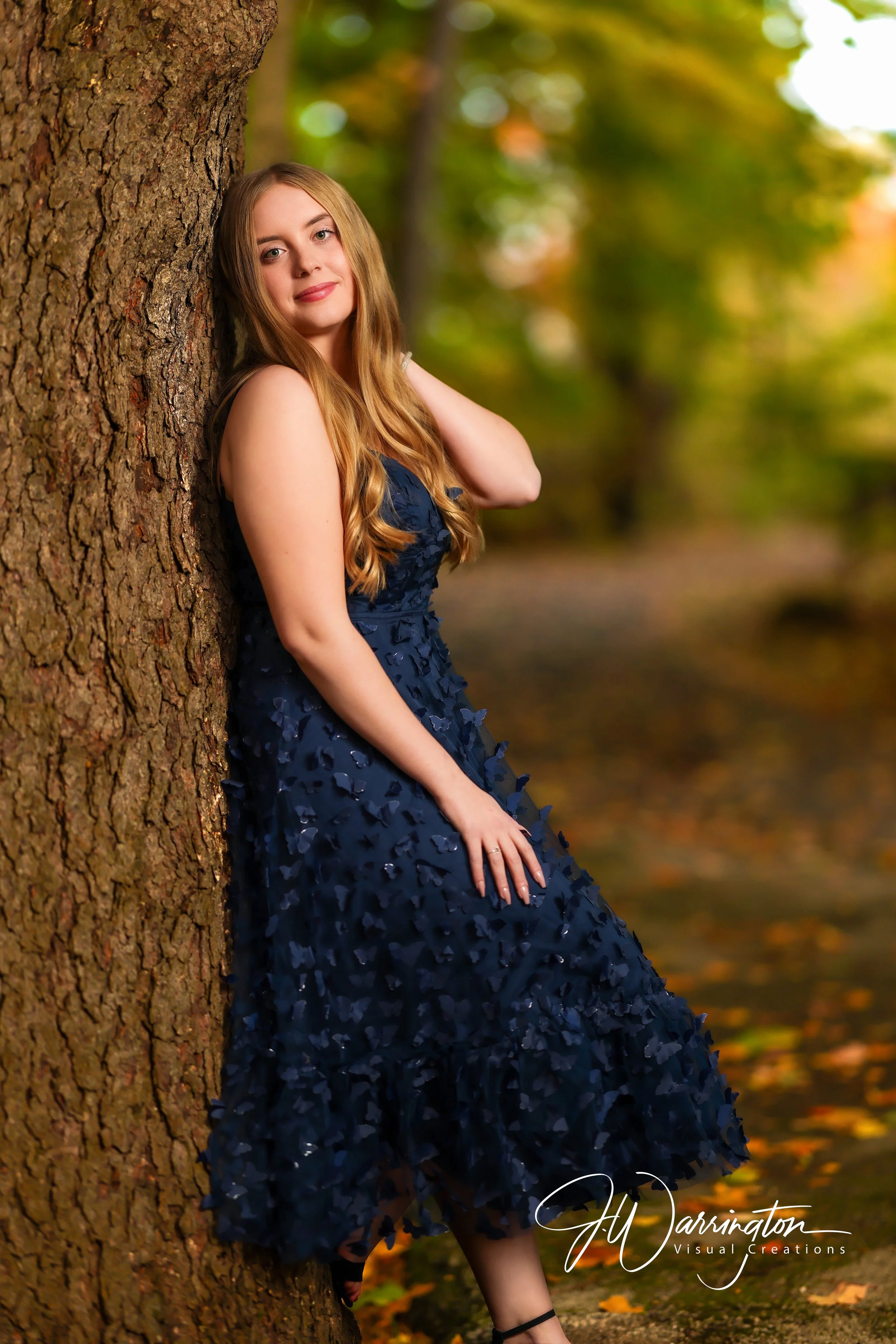 High school senior girl posing gracefully in a long dark navy blue dress while leaning against a tree trunk