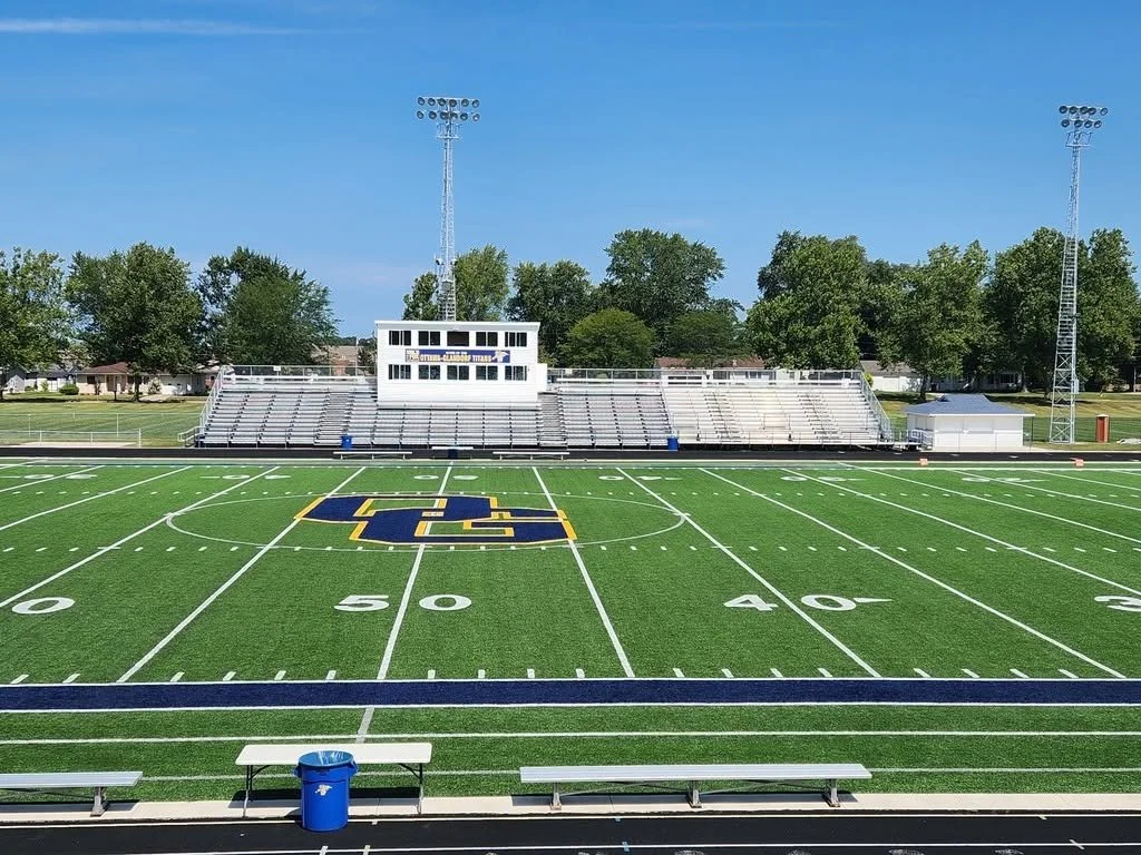 View of an empty football field with a logo at the center, surrounded by bleachers, with tall light towers and green trees in the background.