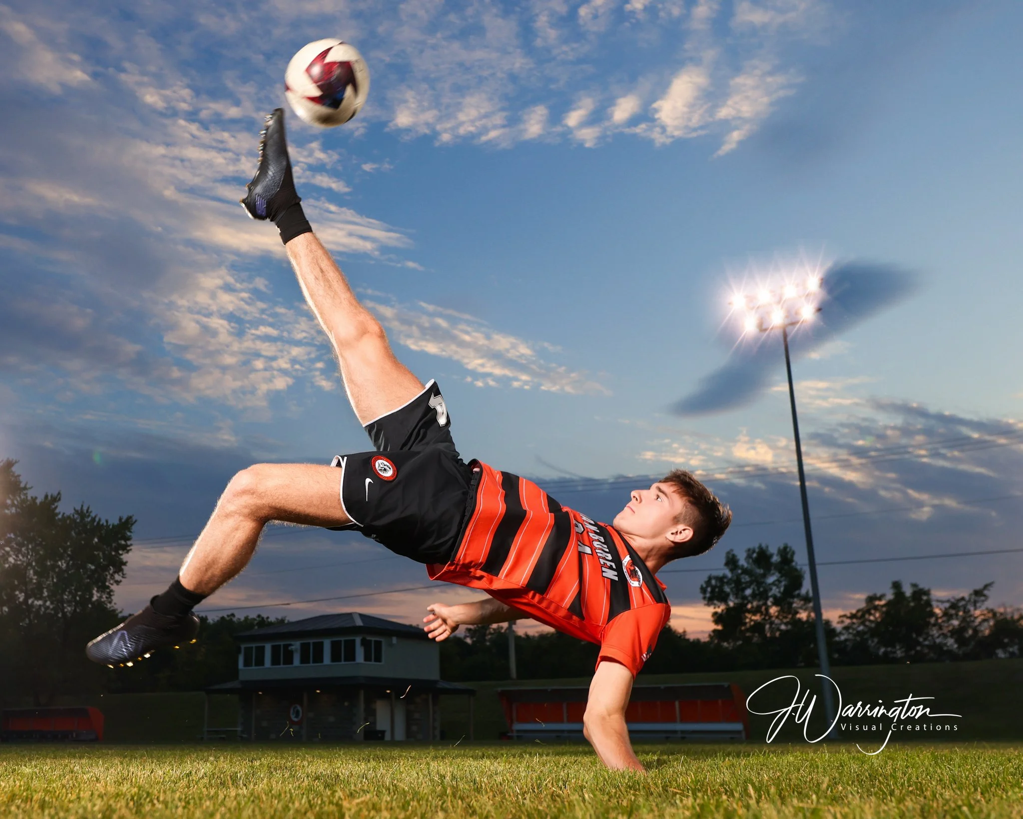 High school senior boy performing a dynamic bicycle kick with a soccer ball under stadium lights during a sunset sports portrait session by Findlay Ohio photographer.
