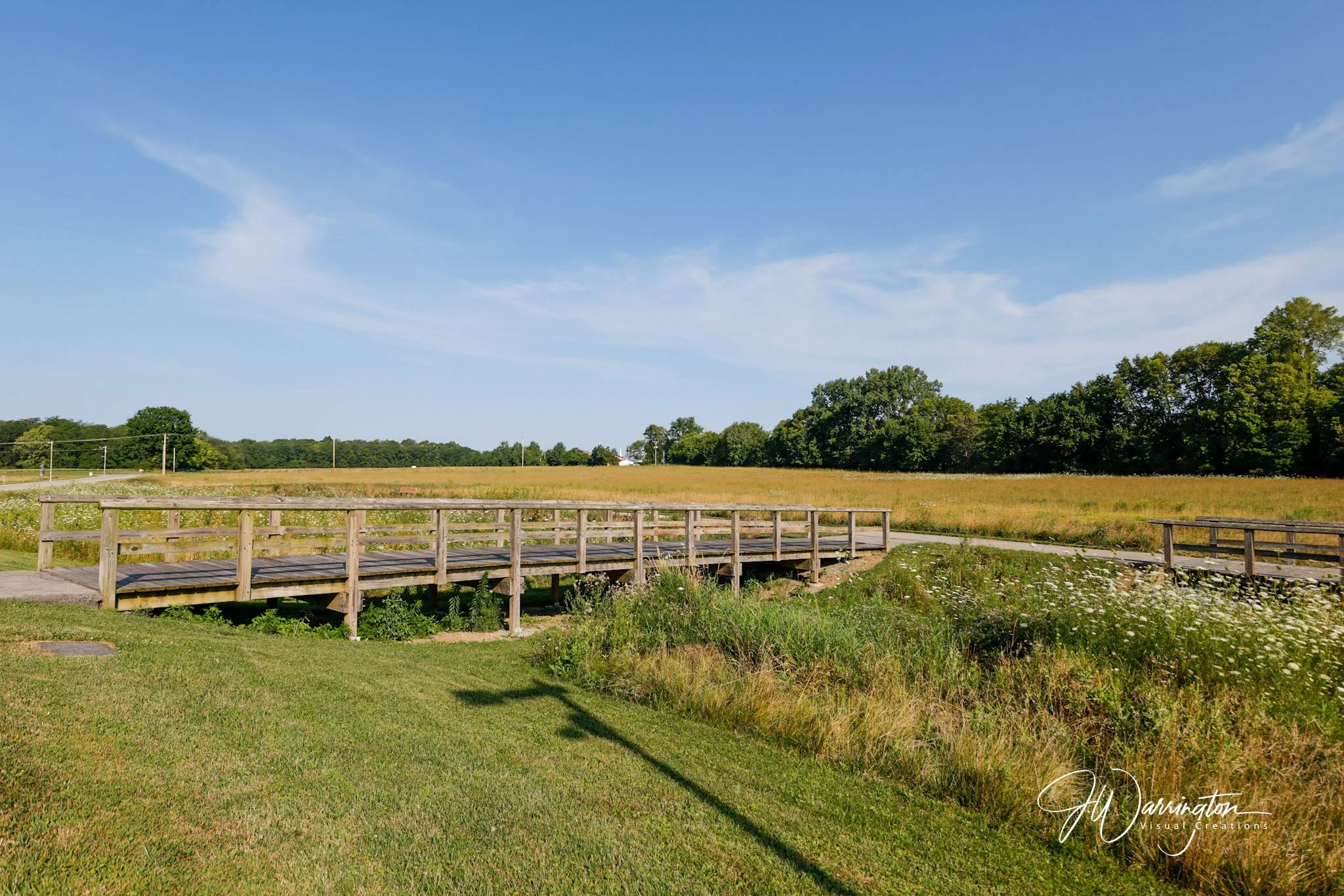 Open grassy field with trees on the horizon at Lauer Historical Farm, wooden footbridge in foreground, under a bright blue sky with some clouds.