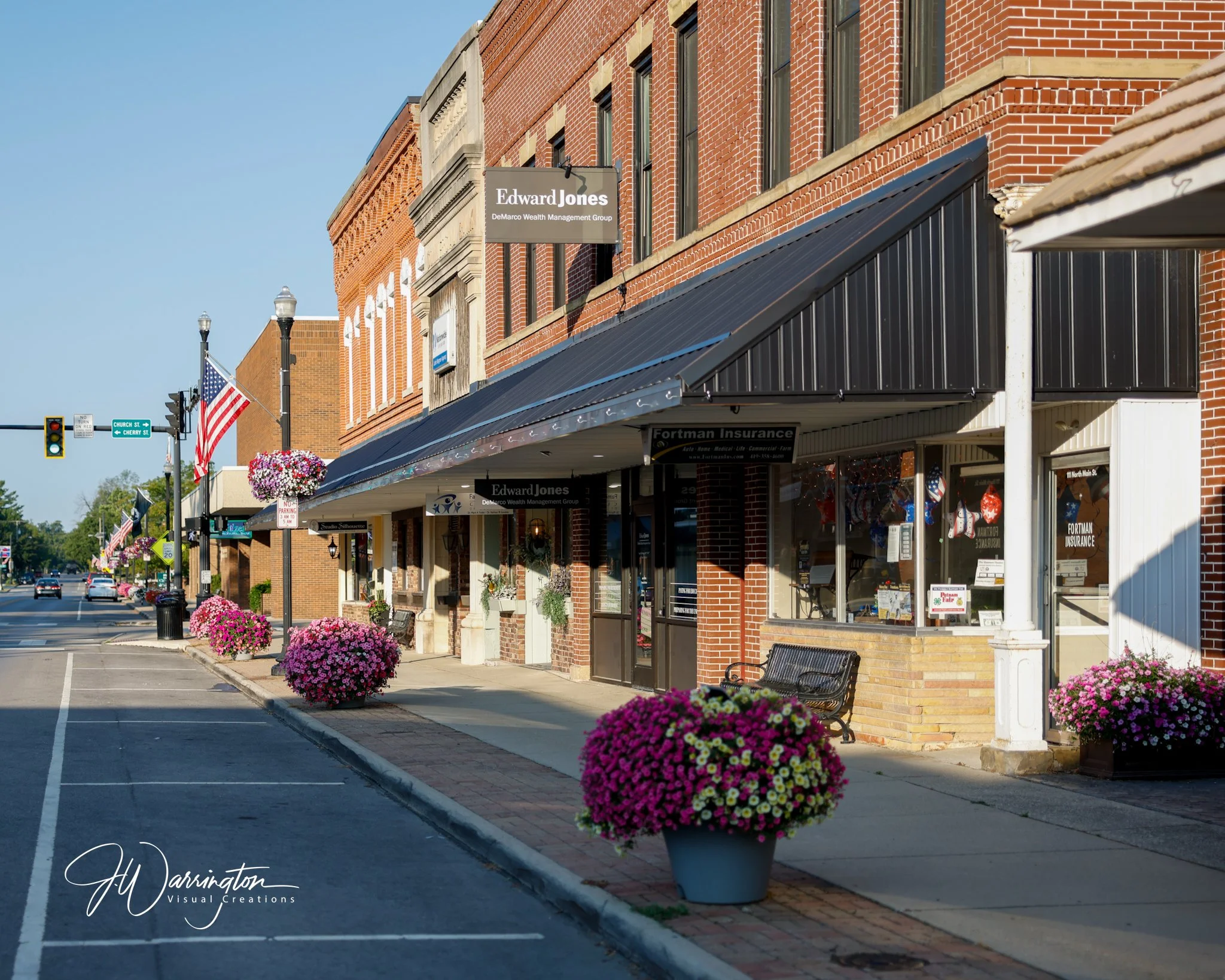 A row of small businesses with brick facades along a clean sidewalk in Historic Downtown Bluffton, decorated with vibrant pink and purple flowers in pots, and American flags hanging outside under clear blue skies.