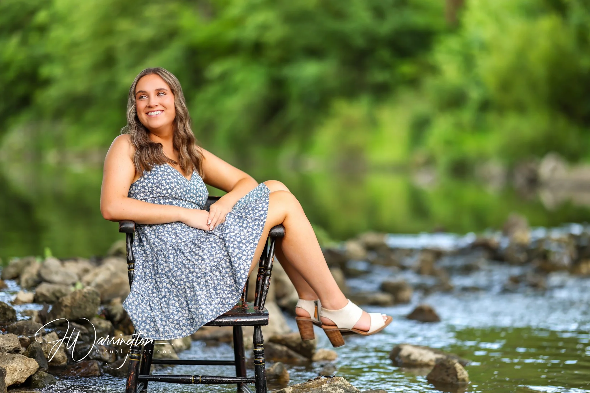 High school senior girl smiling in a blue patterned dress while sitting on a wooden chair by a scenic riverbank