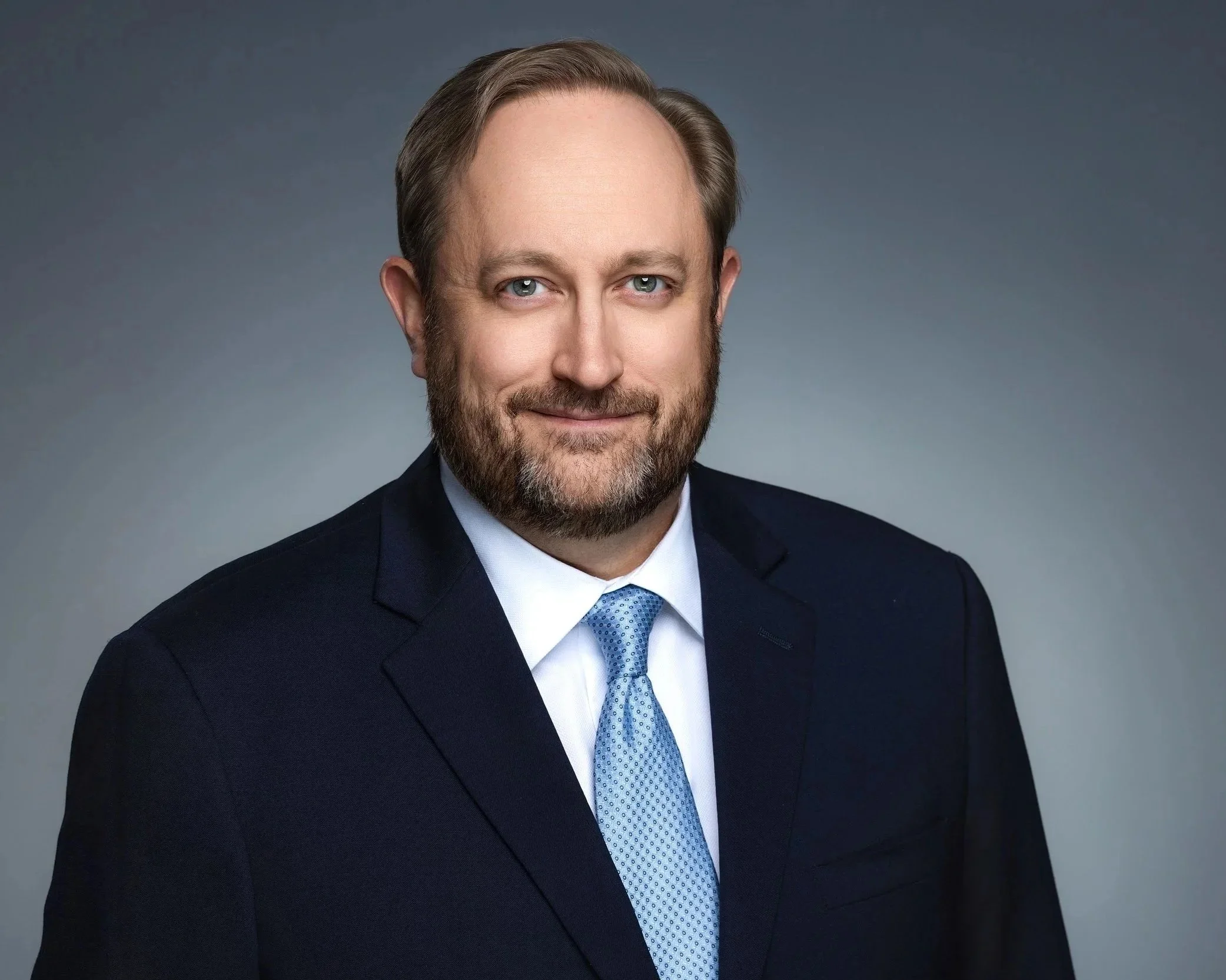 Headshot of a middle-aged man with light skin, brown hair, and a beard, wearing a dark suit, light blue shirt, and patterned tie, against a plain grey background.