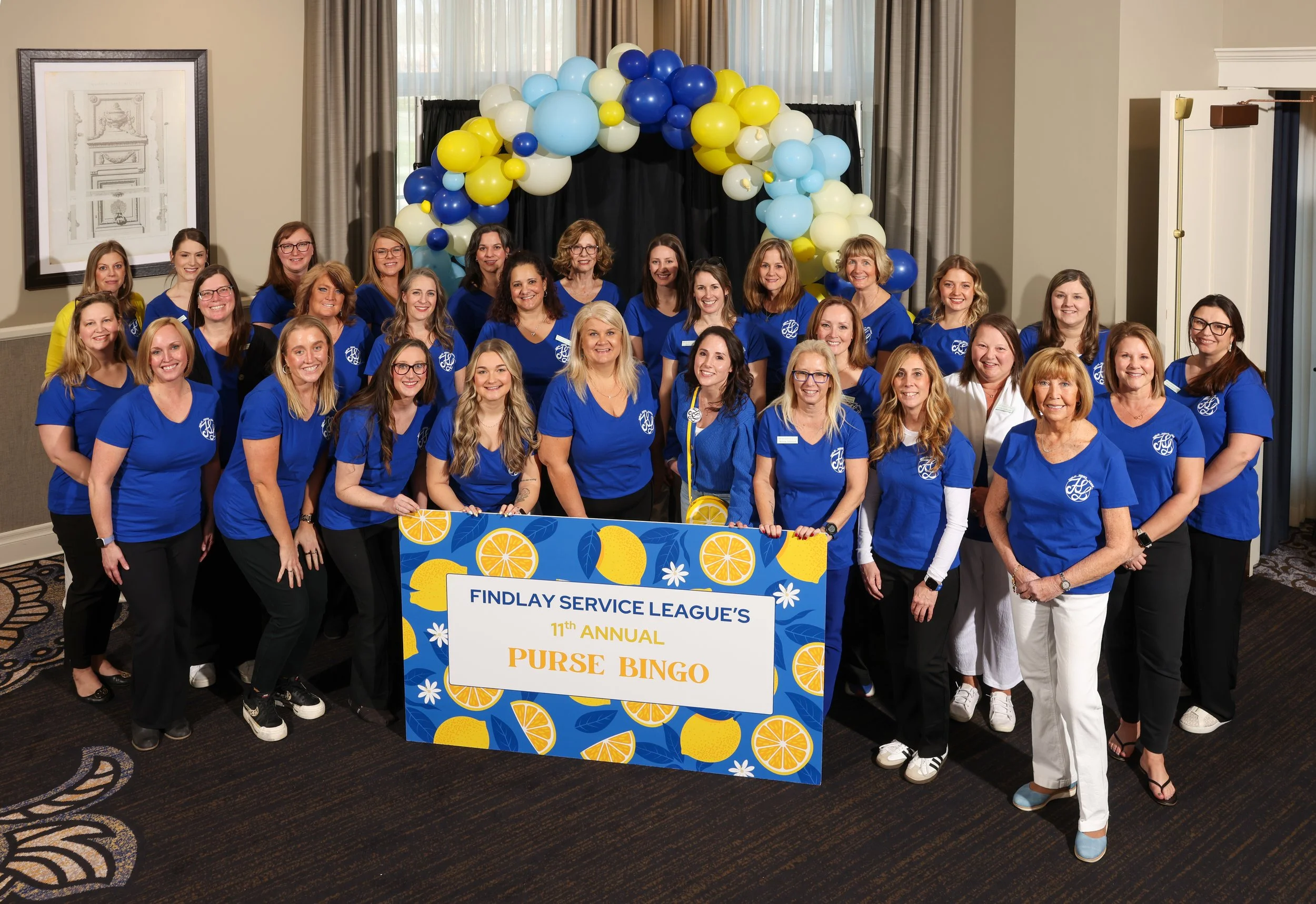 A large group of women dressed in matching blue shirts pose together at an indoor event with a decorative balloon arch captured by Northwest Ohio photographer