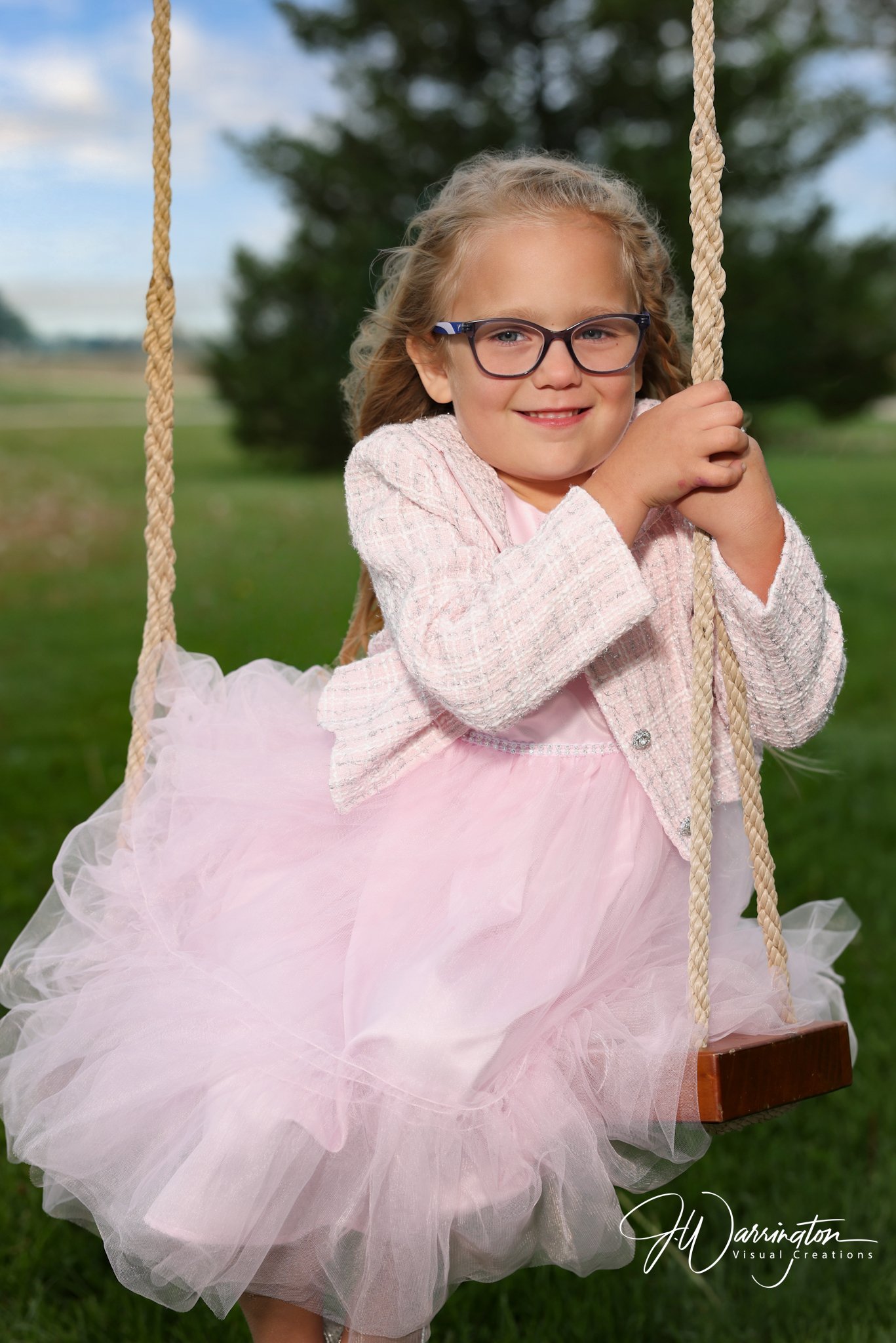 Adorable portrait of a smiling little girl with glasses wearing a white jacket and a pink tulle skirt on an outdoor swing, captured by northwest ohio photographer 