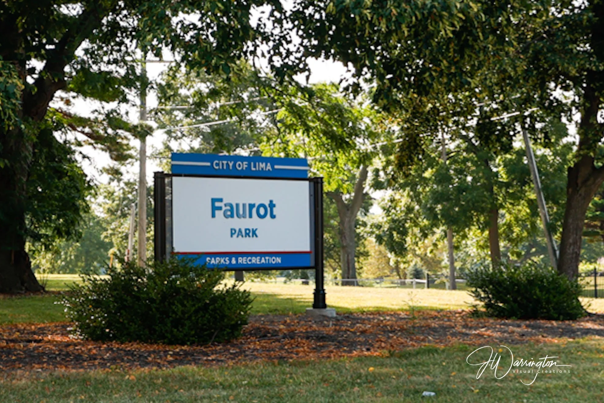 Entrance sign for Faurot Park in the city of Lima, surrounded by green trees and foliage, with a walking path and grass in the background.