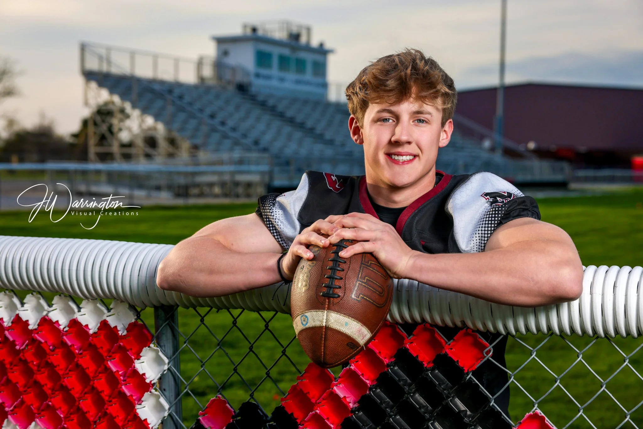 High school senior male leaning on fence with press-box behind him during a sunset.