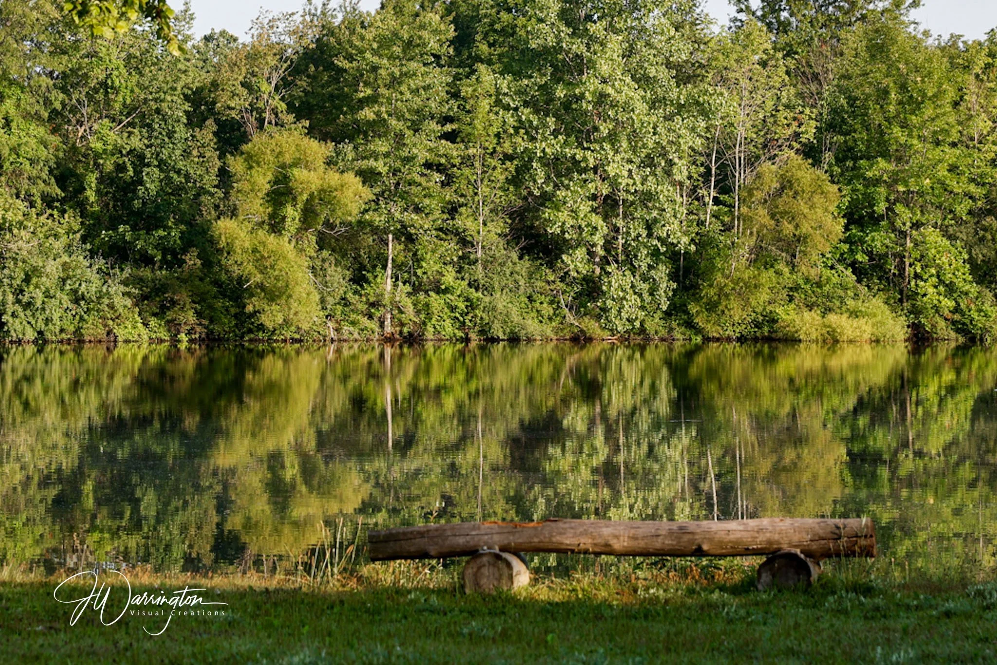A serene scene at Nature Preserve, in Bluffton, Ohio, green trees along a riverbank, with their reflection mirrored in the still water, and a fallen log in the foreground.