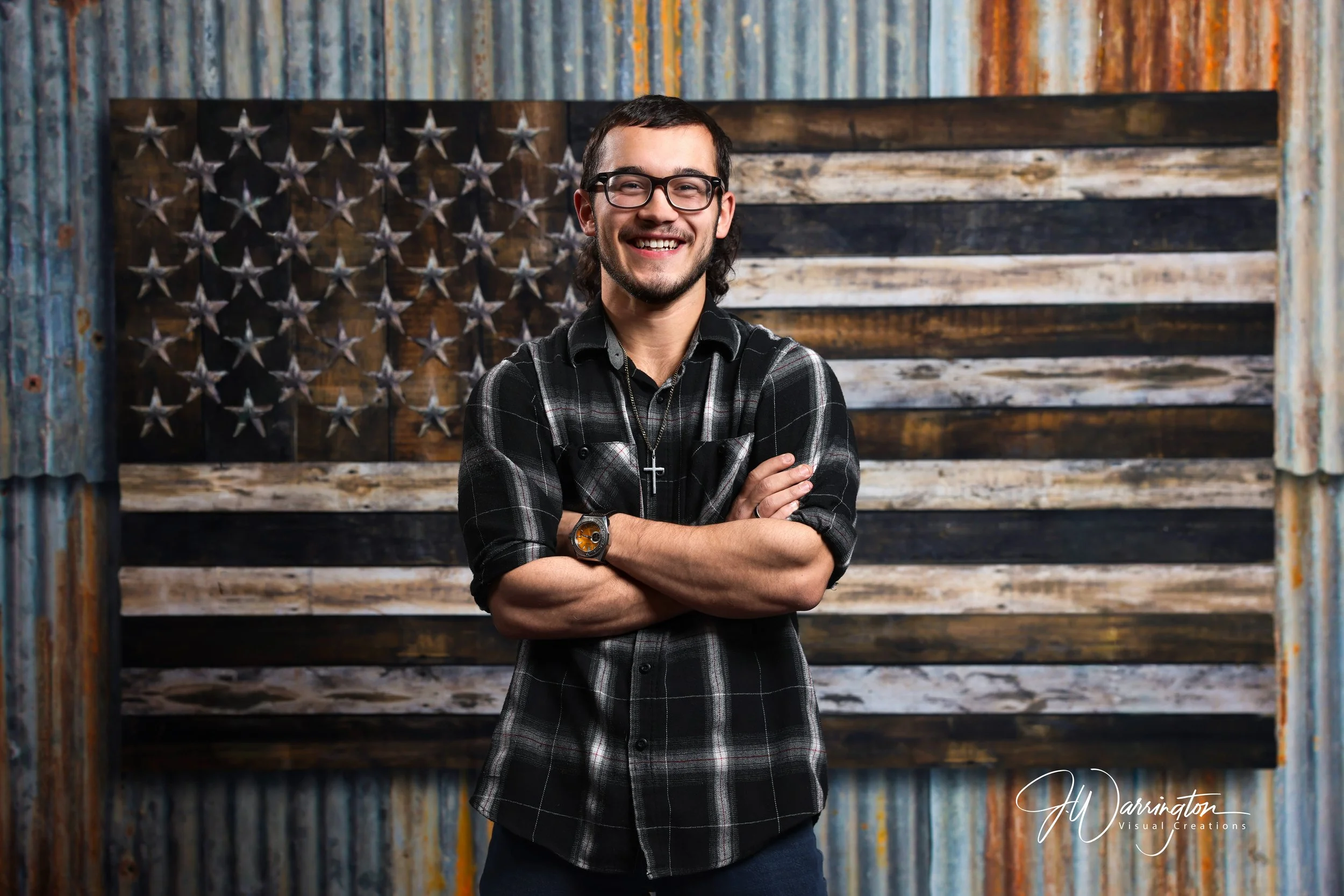 A young man with glasses and a beard smiling, standing with crossed arms in front of a rustic American flag made of wood and metal stars.