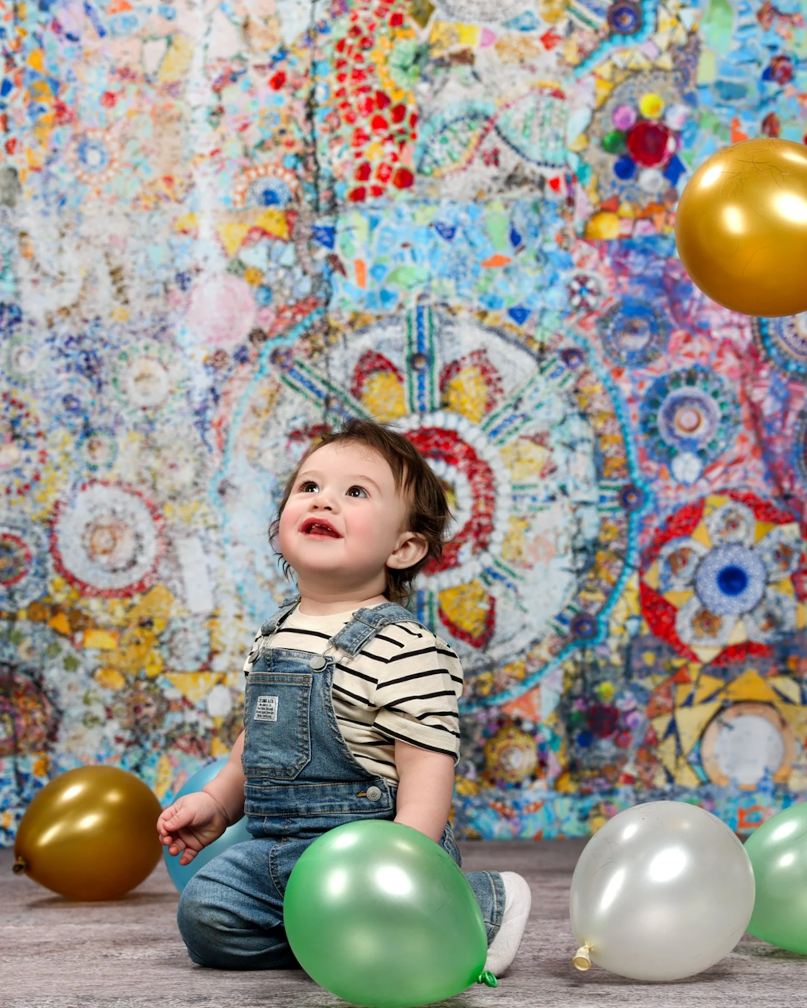 Birthday milestone studio portrait of a smiling baby boy wearing overalls, sitting among colorful balloons in front of a vibrant, artistic background, captured by northwest ohio photographer 