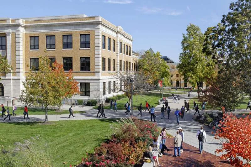 Campus scene with students walking along pathways and benches, surrounded by trees and a large academic building in the background.
