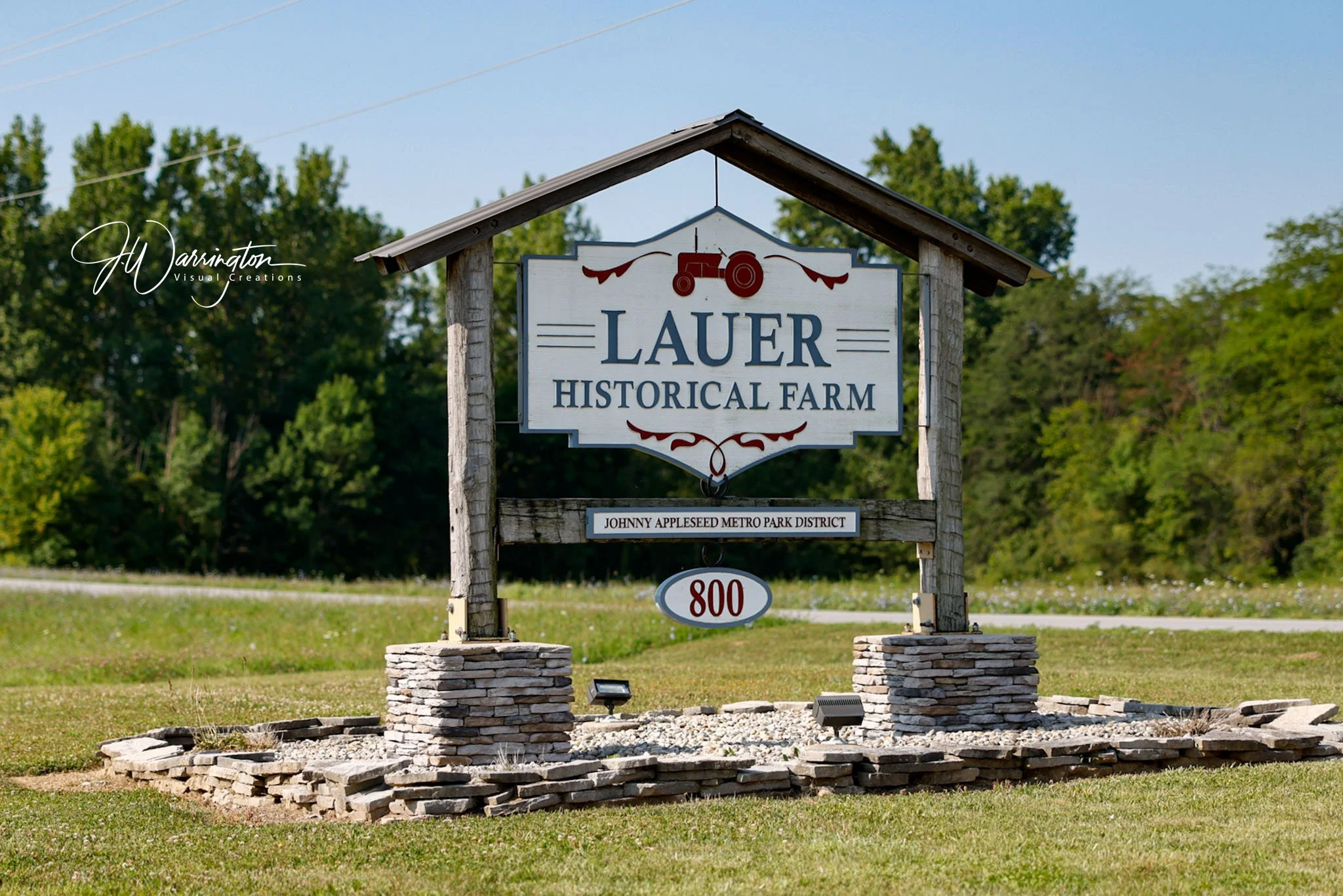 A sign for the Lauer Historical Farm at Johnny Appleseed Metro Park District, featuring a red tractor illustration and surrounded by greenery and trees.