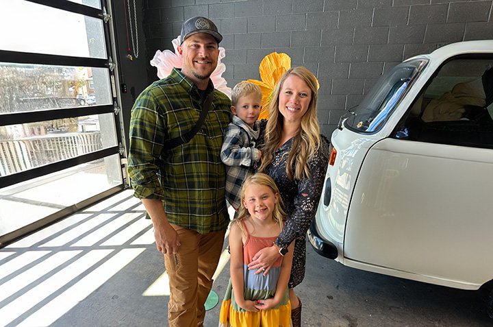 A family of four posing indoors next to a vintage white car. The father is wearing a green plaid shirt and cap, holding a young boy. The mother, in a patterned dress, stands beside a young girl in a colorful dress. The background features a dark wall and garage door.