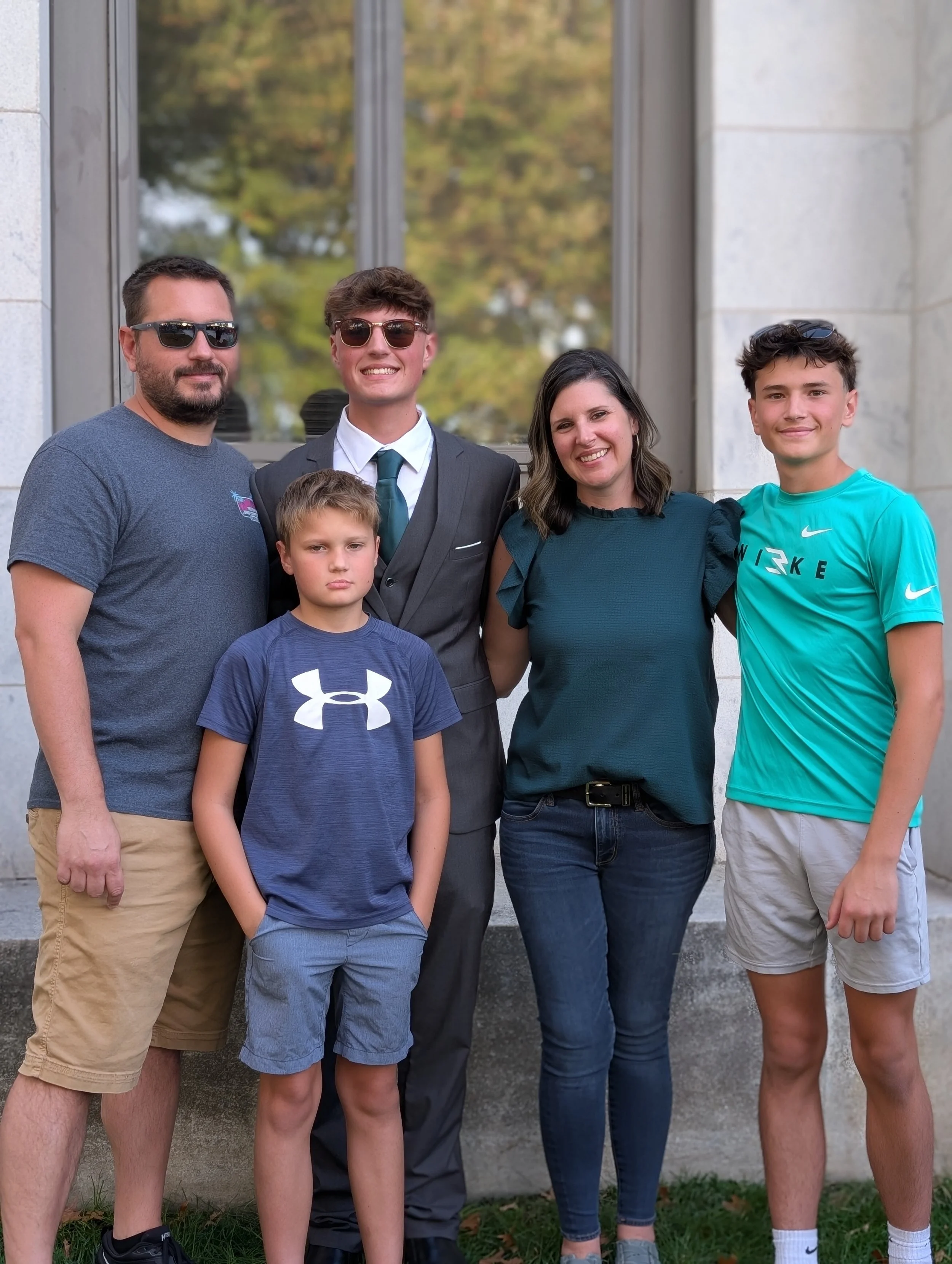 A group of six people, including two adults and four boys, standing outdoors in front of a building. The man on the left is wearing sunglasses, a gray T-shirt, and tan shorts. The boy in front of him has short blond hair, a blue Under Armour T-shirt, and gray shorts. The young man in the center is wearing sunglasses, a dark suit, white shirt, and a teal tie. The woman next to him has shoulder-length dark hair, a dark teal blouse, and jeans. The boy on the right is wearing a bright teal Nike T-shirt and beige shorts. They are smiling and standing close together.