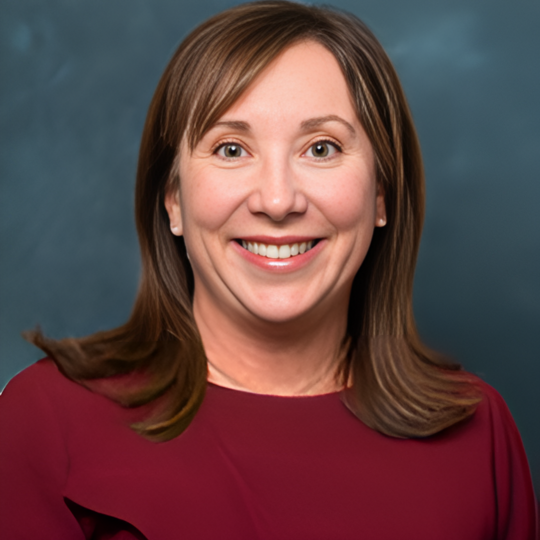 A woman with shoulder-length brown hair smiling at the camera, wearing a maroon top, against a blue background.
