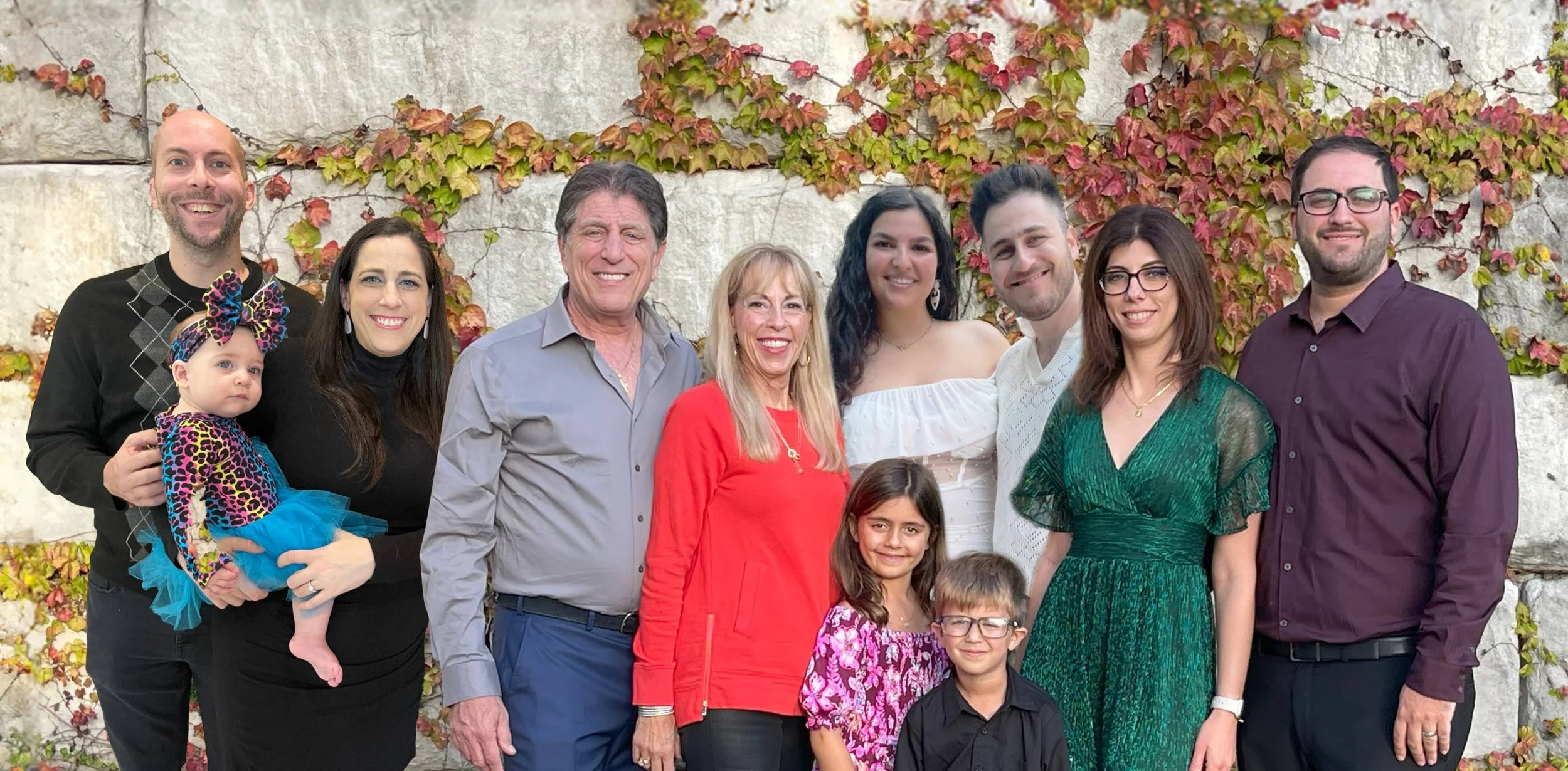 A group of ten people, including three children, smiling outdoors in front of a stone wall with colorful autumn leaves.