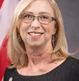 A smiling woman with glasses and shoulder-length blonde hair, wearing a black top, in an indoor setting.