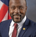 A smiling African American man in a navy suit, white shirt, and red tie, standing in front of an American flag.