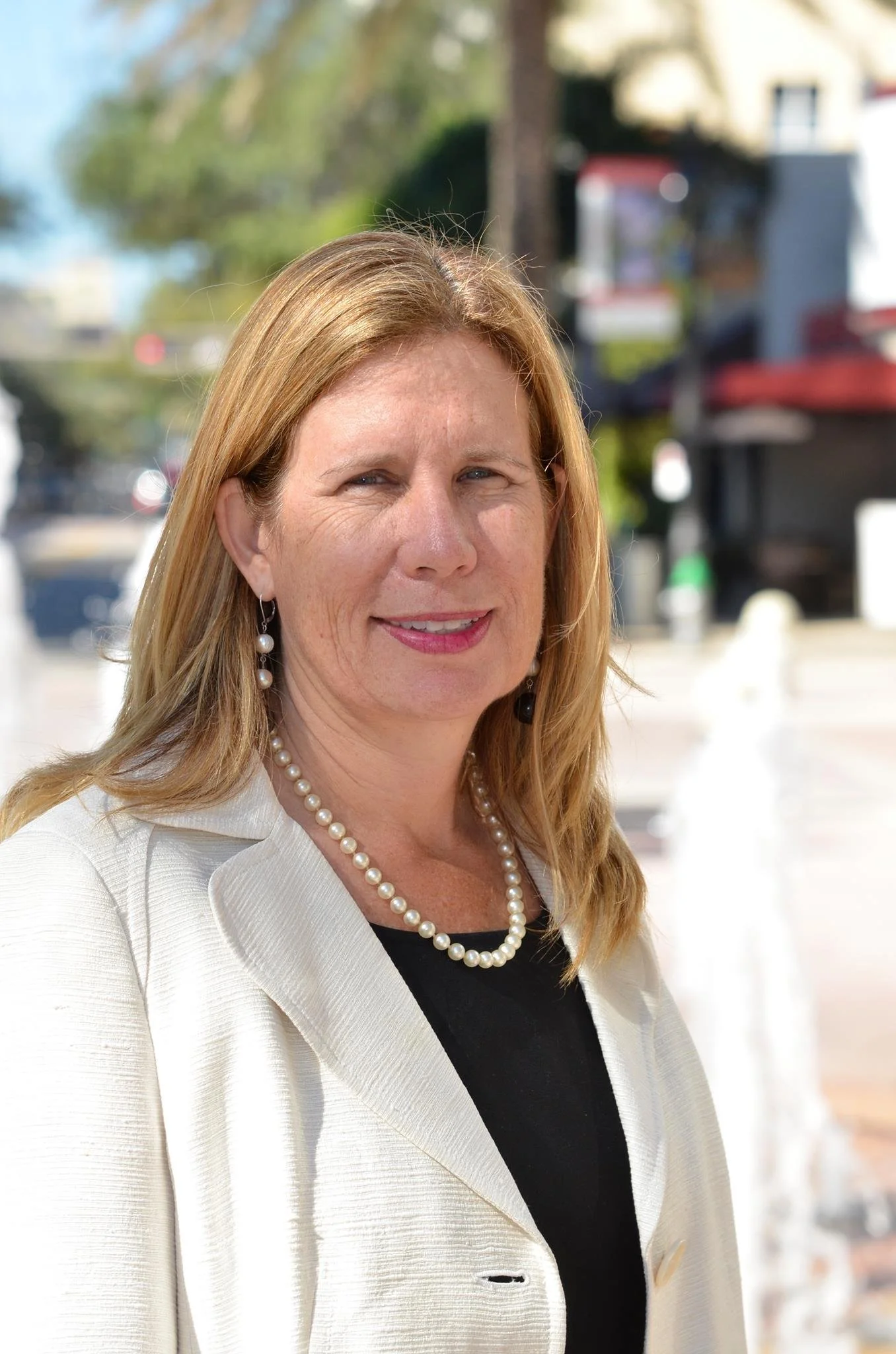 A woman with shoulder-length blonde hair wearing a white blazer, black top, pearl necklace, and pearl earrings standing outdoors on a sunny day with trees and blurred buildings in the background.