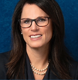 A woman with long dark hair wearing glasses, a dark blazer, and a pearl necklace, smiling against a dark blue background.