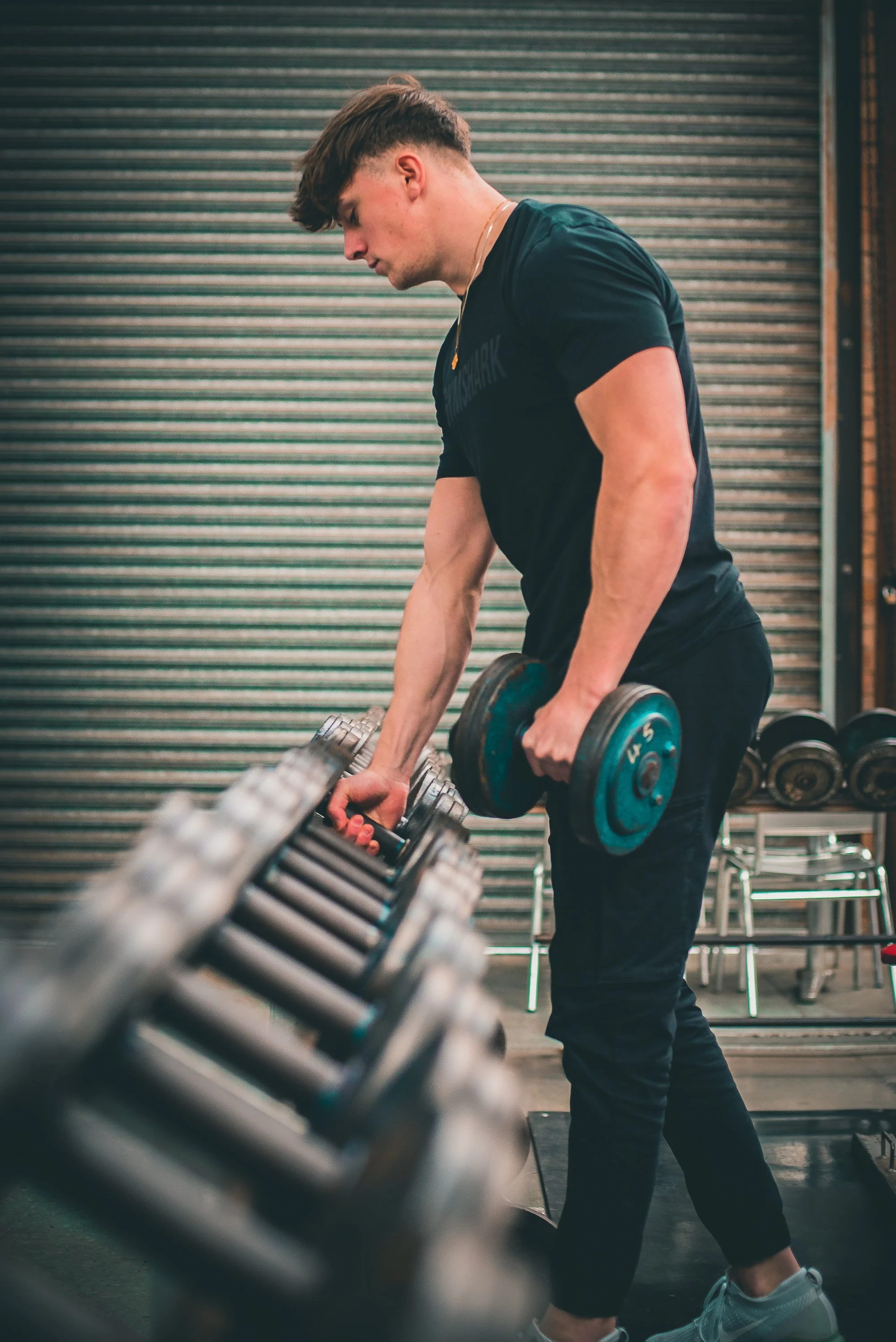A man lifting a dumbbell in a gym, standing next to a rack of dumbbells and metal chairs with a roll-up door in the background.