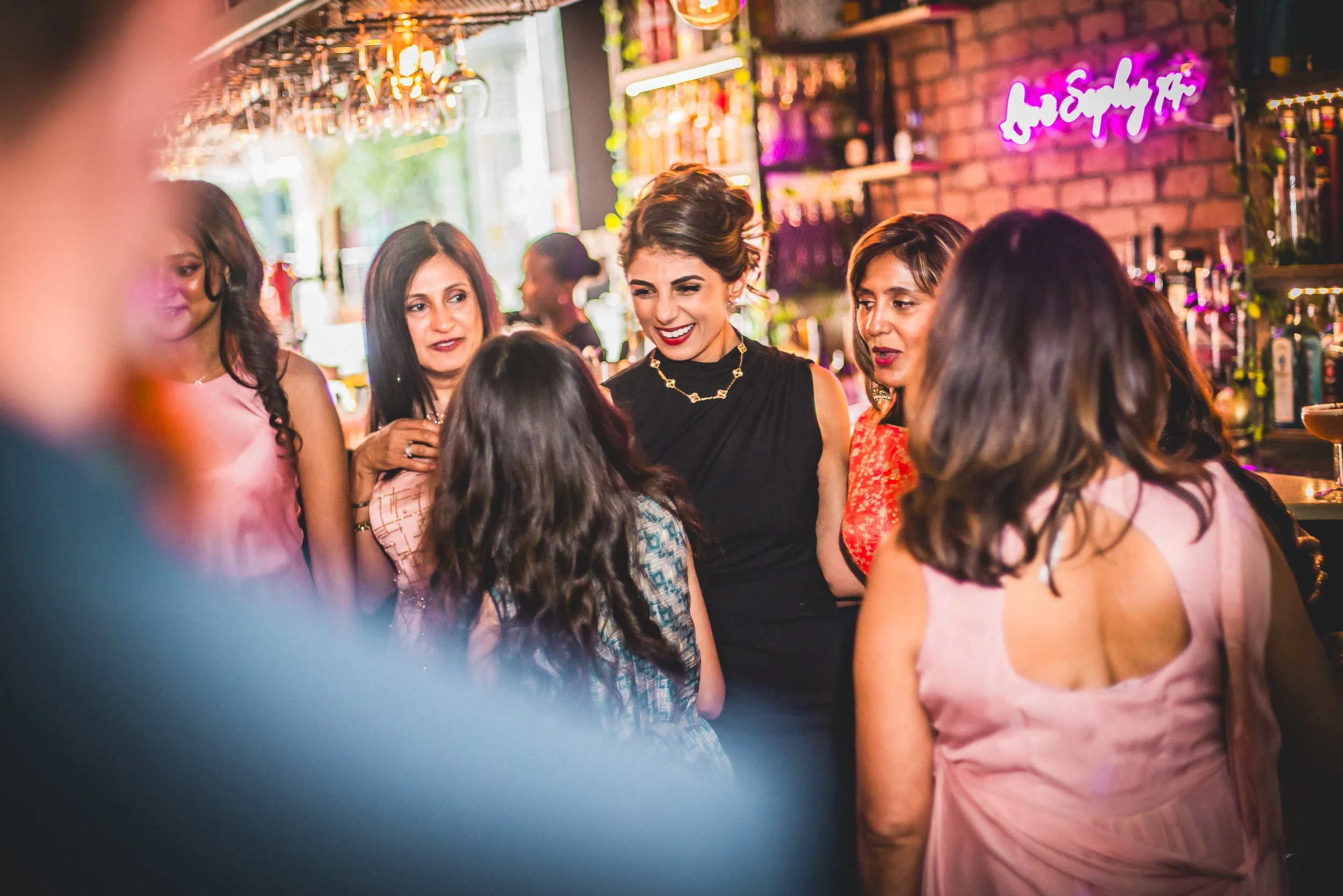 A group of women socializing in a cozy bar with exposed brick walls, purple neon sign, and hanging wine glasses.