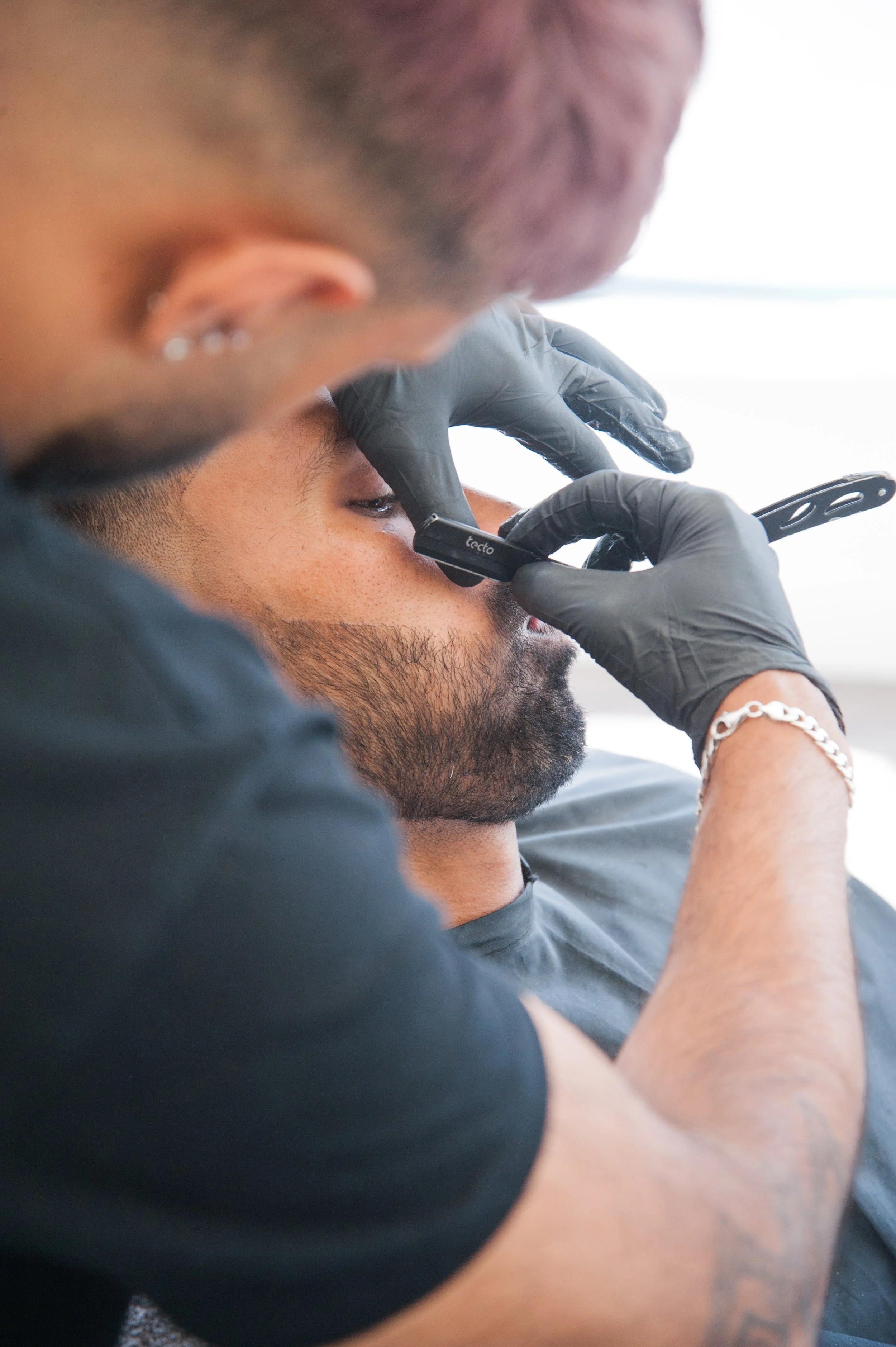 A person with a beard and tattoos gets a nose piercing from a professional piercer. The piercer is wearing black gloves and holding a piercing tool, while the person lies back with eyes closed.