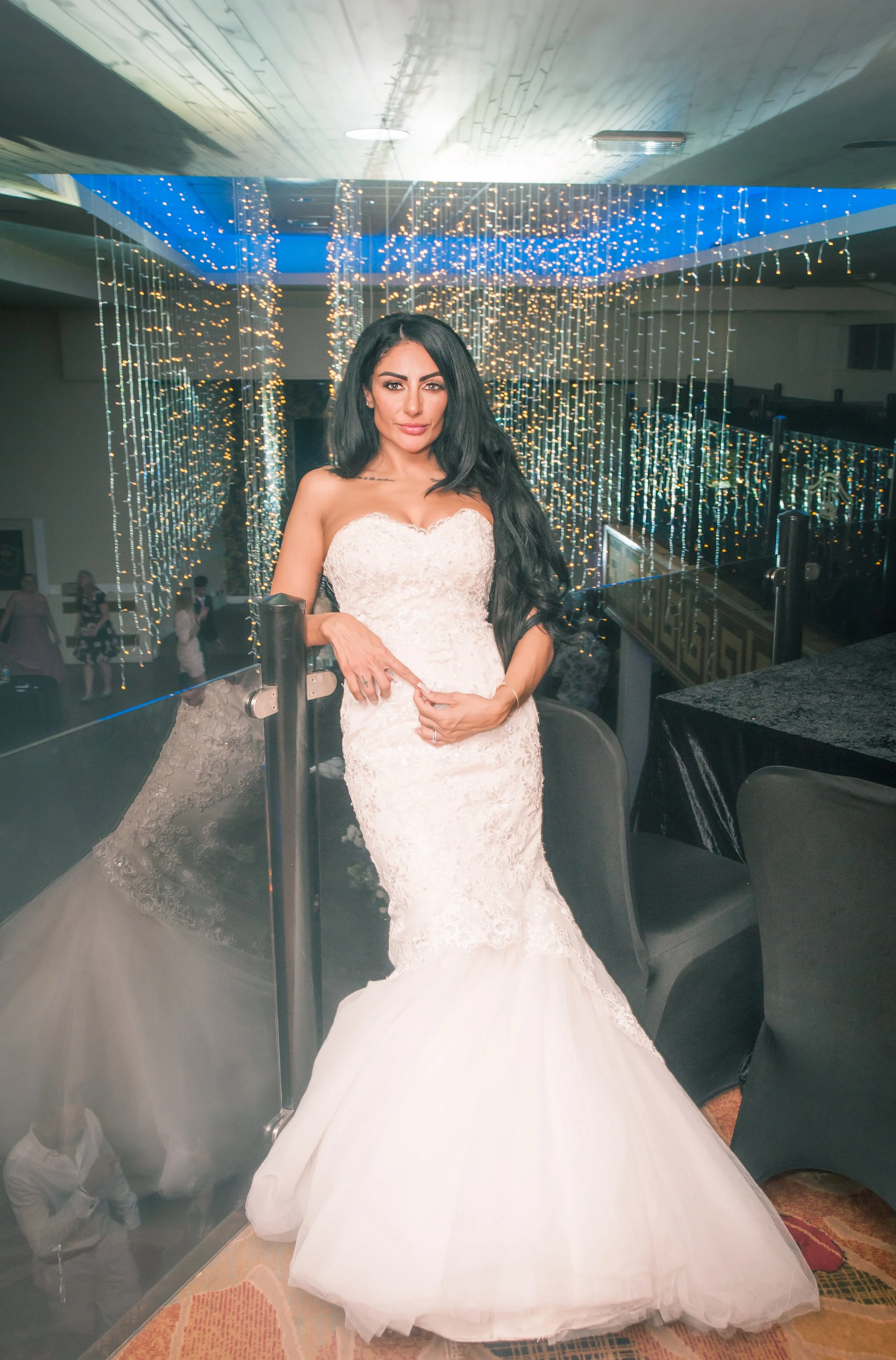 A woman in a white lace wedding dress standing next to a glass panel with string lights in the background, in an indoor venue.