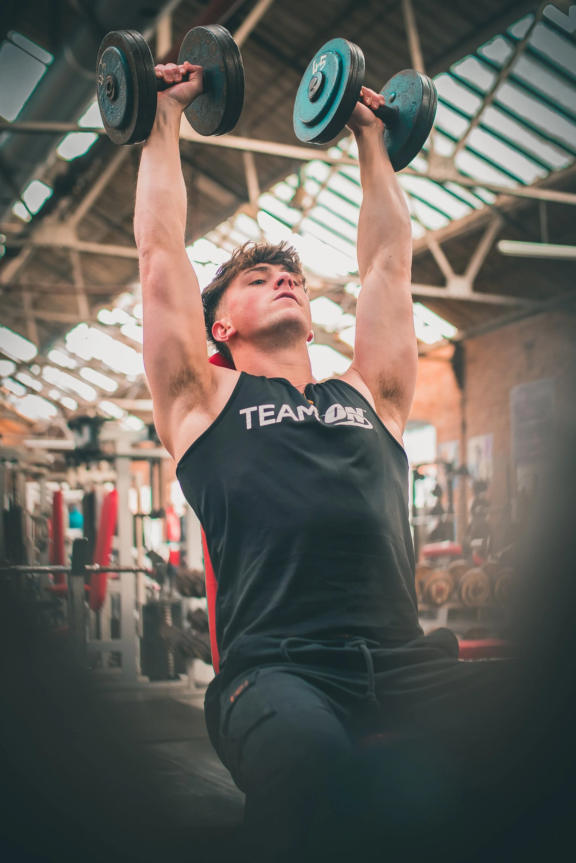 A young man in a gym lifting dumbbells overhead during a workout.