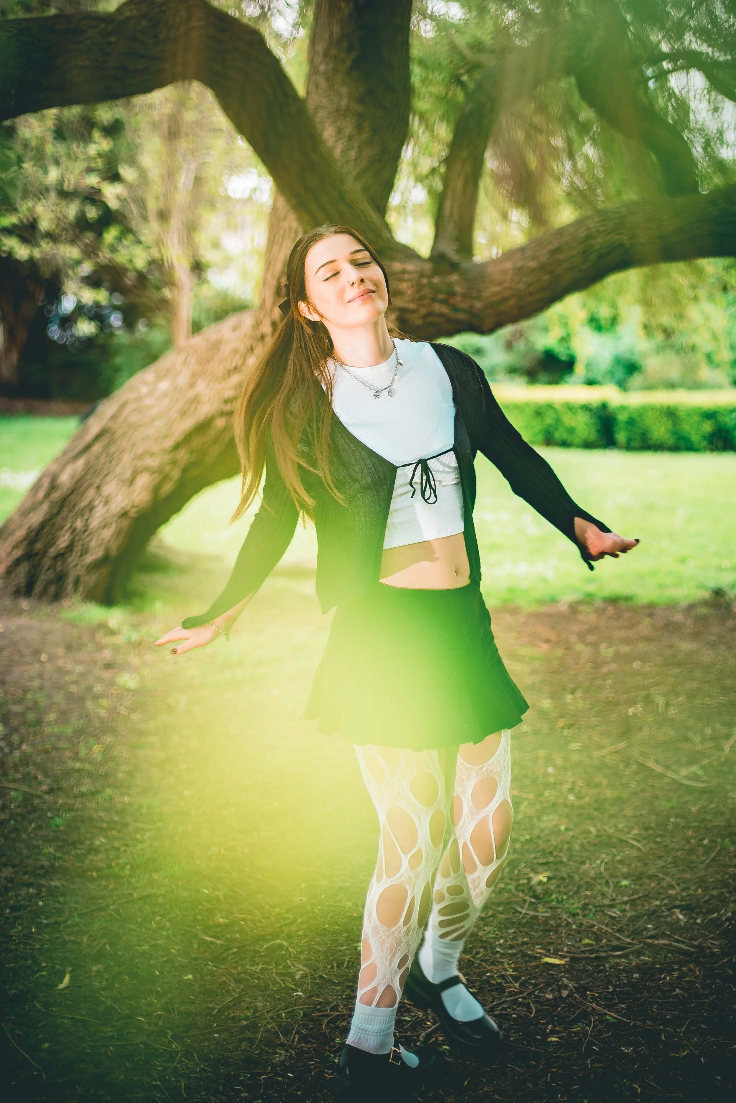 Young woman in a black jacket, white crop top, and torn leggings standing with arms open outdoors near a large tree in a park with sunlight filtering through leaves.
