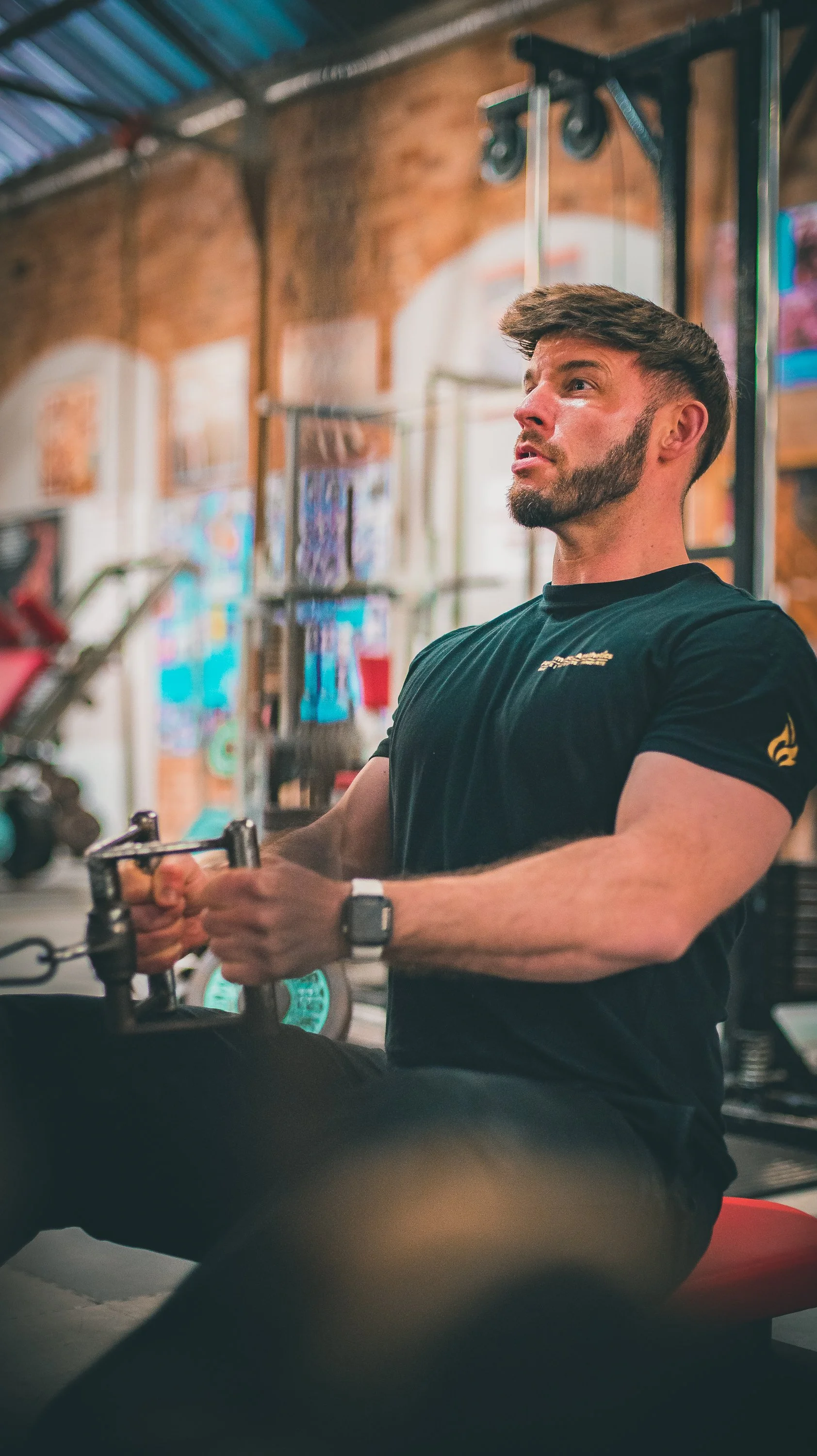 A man working out in a gym, using a rowing machine. He has a beard, short brown hair, and is wearing a black t-shirt and a wristwatch. The gym has exposed brick walls and colorful artwork in the background.